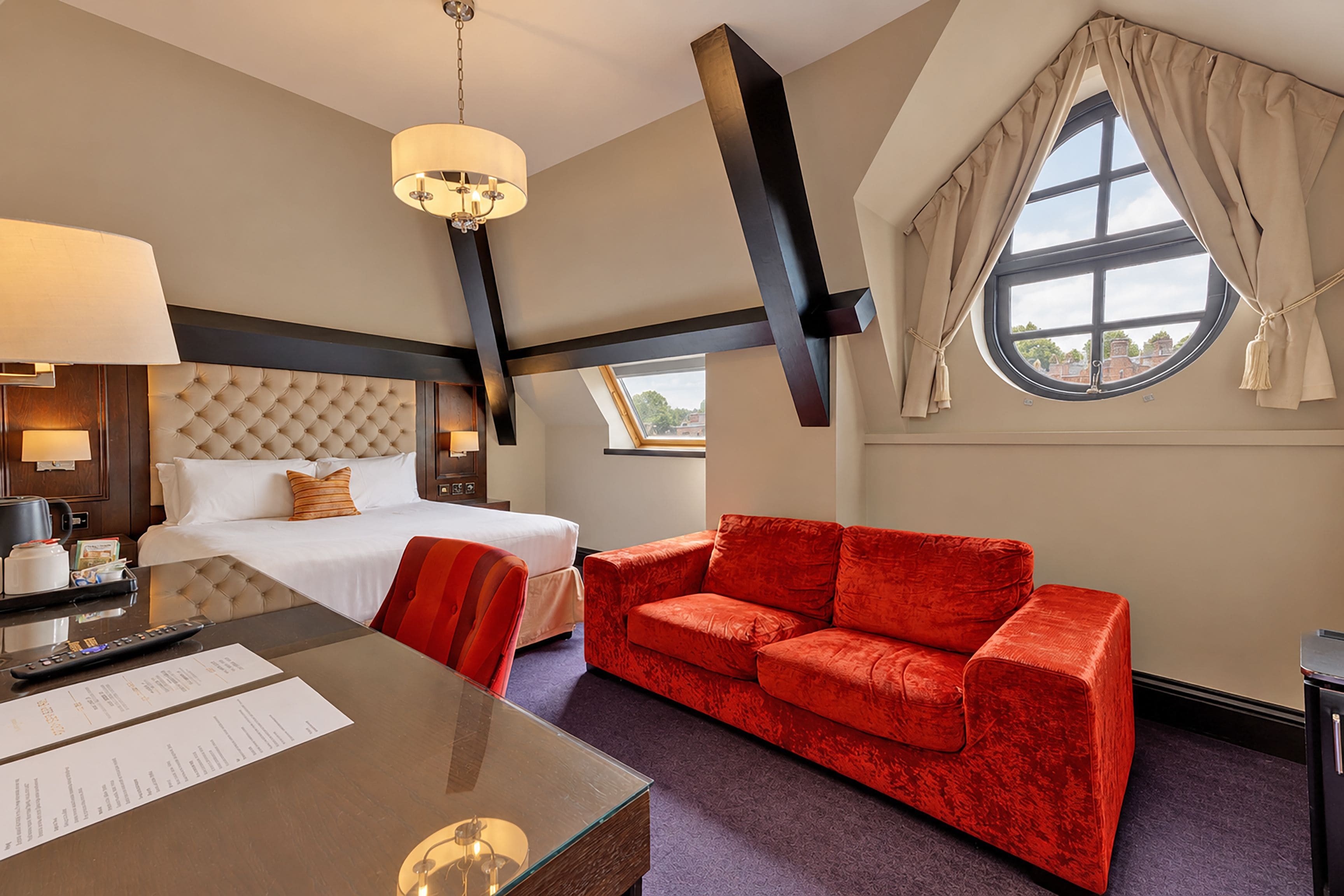 A hotel room with a bed, a red sofa, a desk with a lamp, and a large window with curtains.