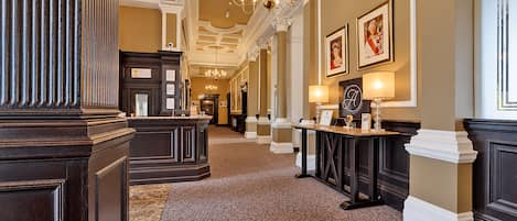 A grand hallway with ornate columns, a patterned carpet, and a chandelier.