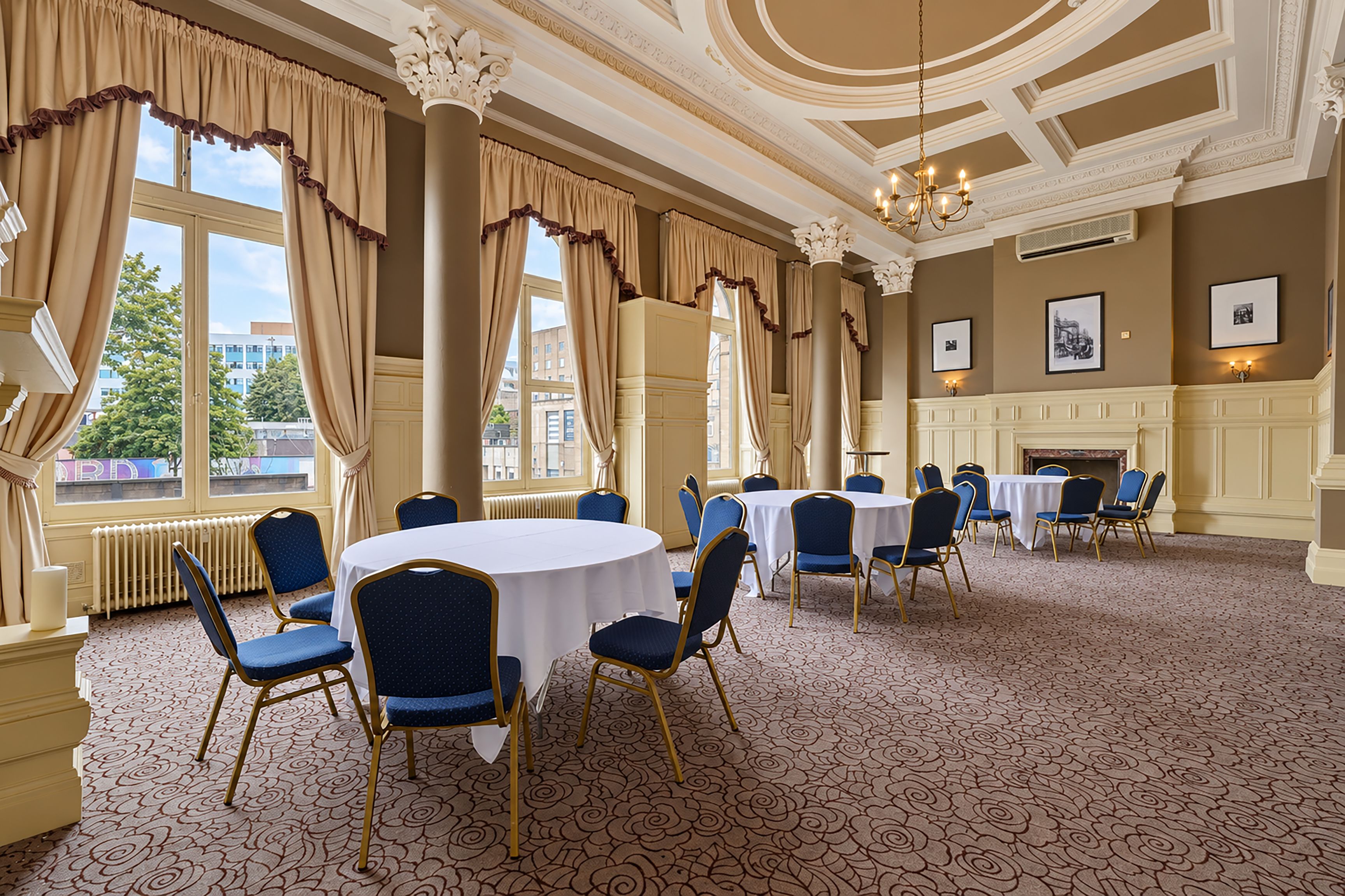 Une salle à manger élégante, avec des tables rondes et des chaises bleues, de grandes fenêtres ornées de rideaux et une cheminée.