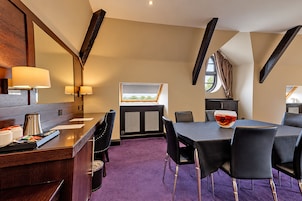 A hotel room with a dining area, a large mirror, a window with curtains, and a view of greenery outside.