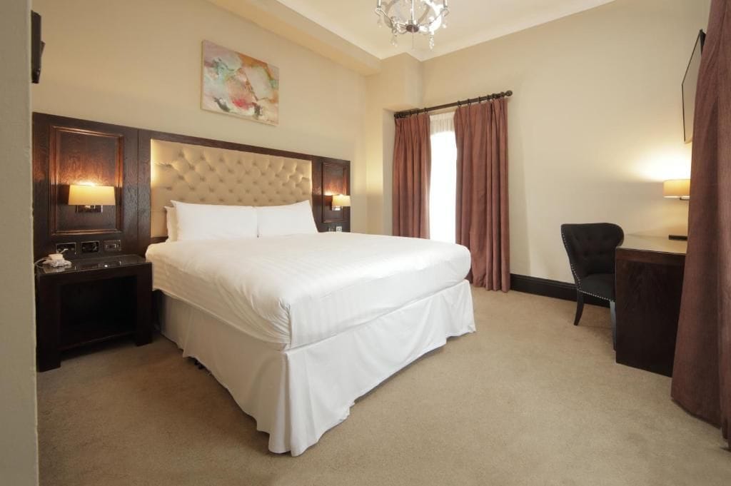 A hotel room with a large bed, a nightstand with a lamp, a chair, and a desk with a lamp.