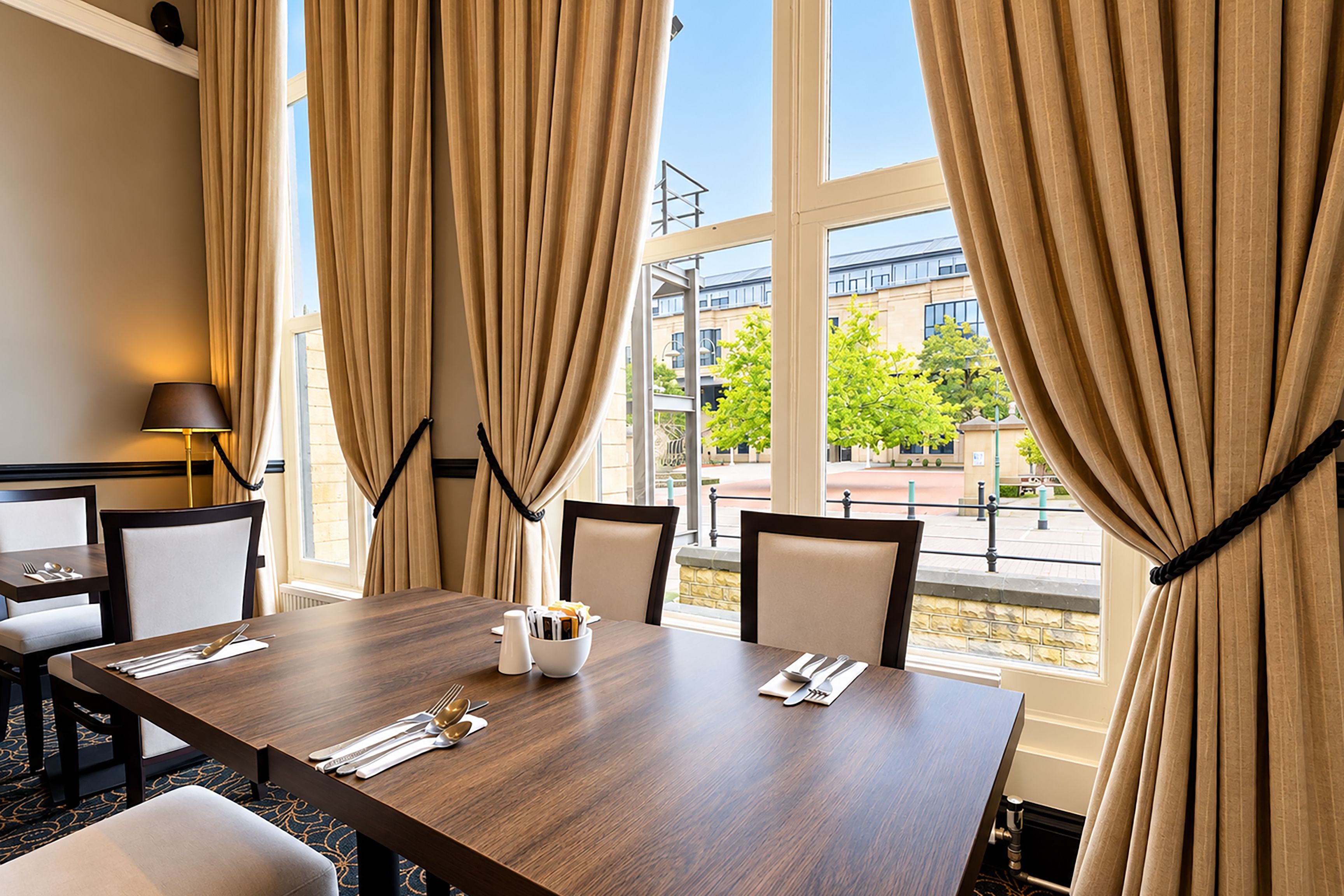 A hotel room with a dining table set for four, two chairs on each side, a lamp, and a view of the outside.