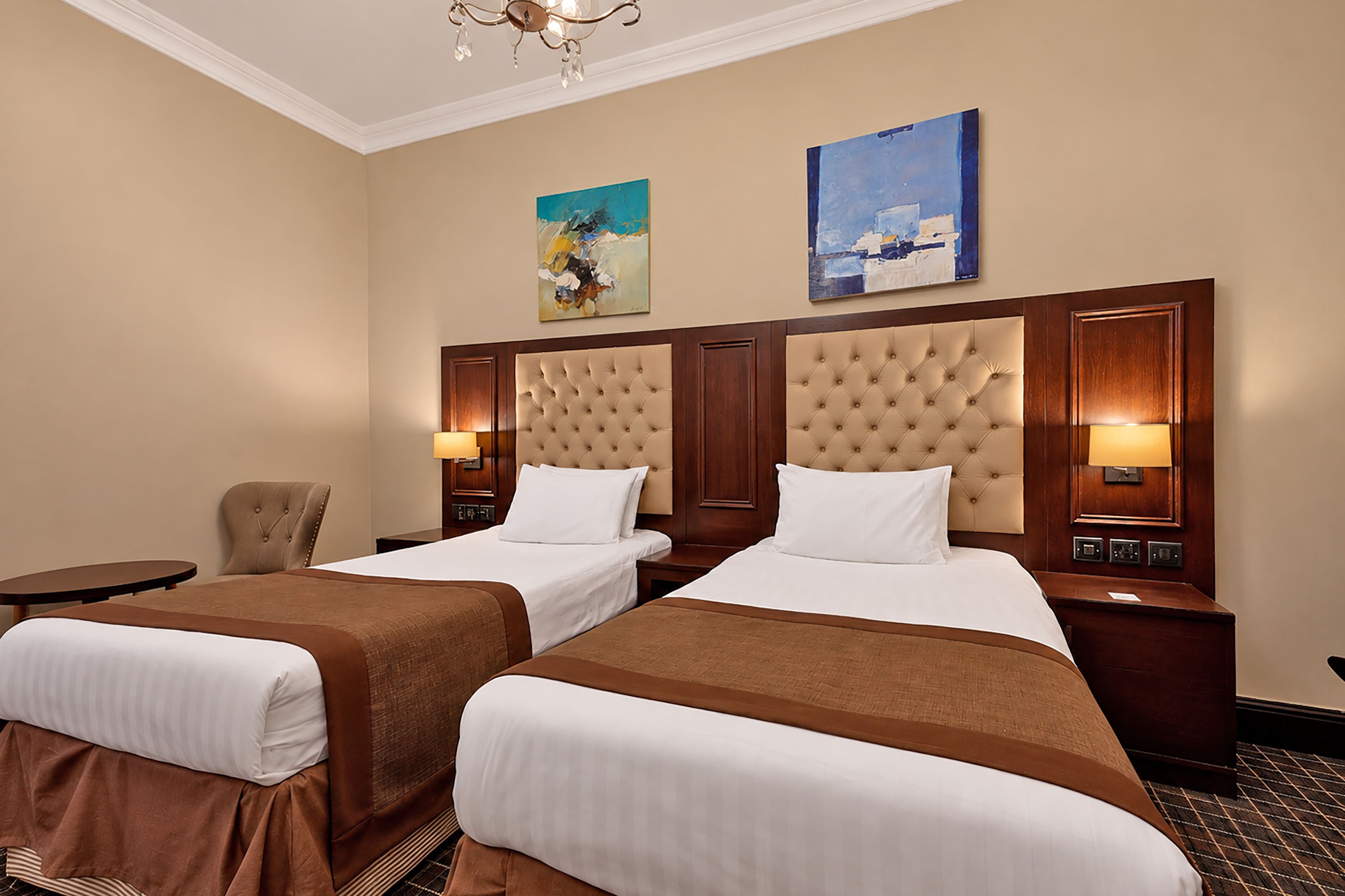 Een hotelkamer met twee bedden, een stoel, een klein tafeltje en kunstwerken aan de muur.