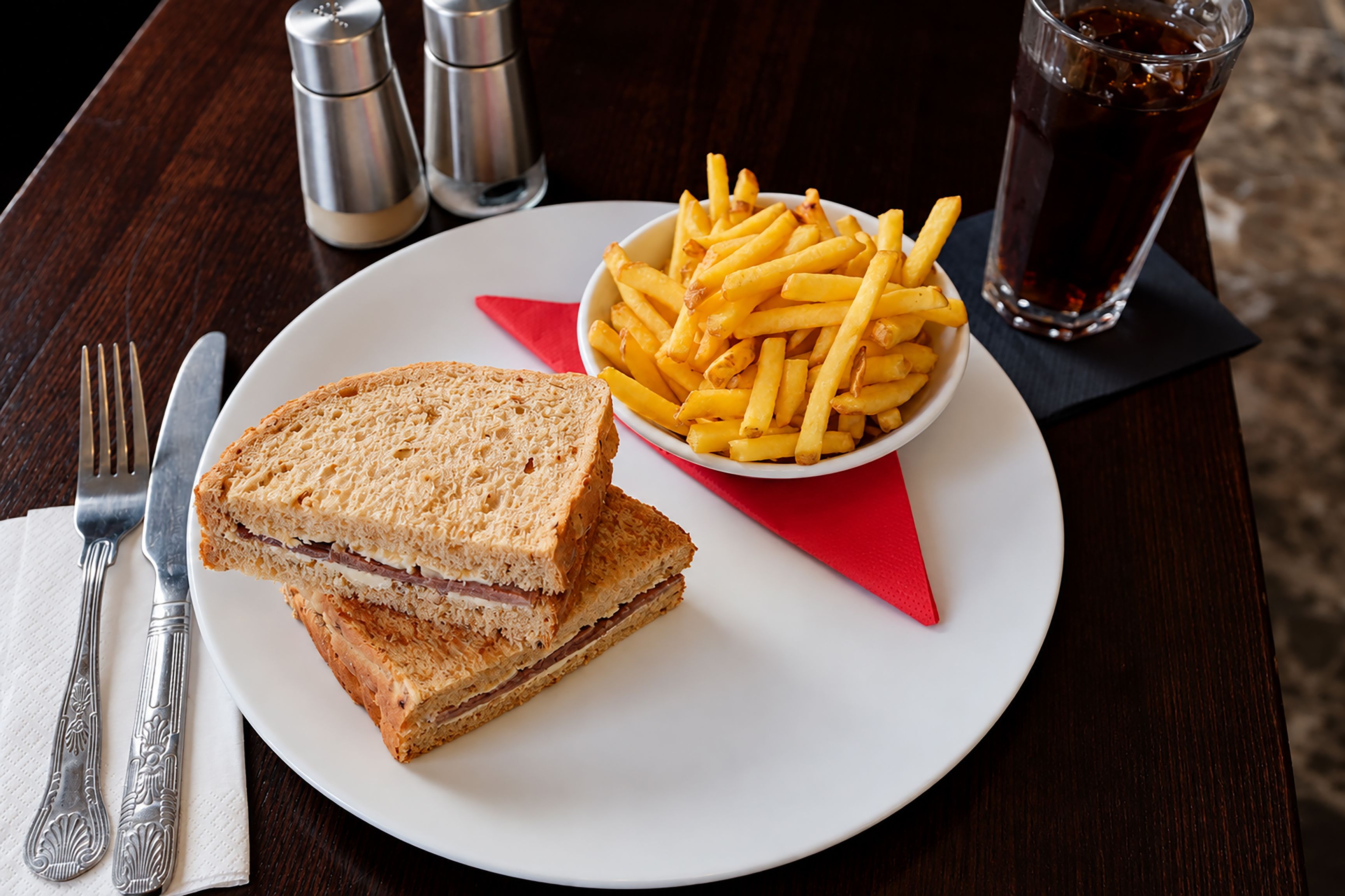 A sandwich with fries on a plate, a glass of soda, and cutlery on a table.