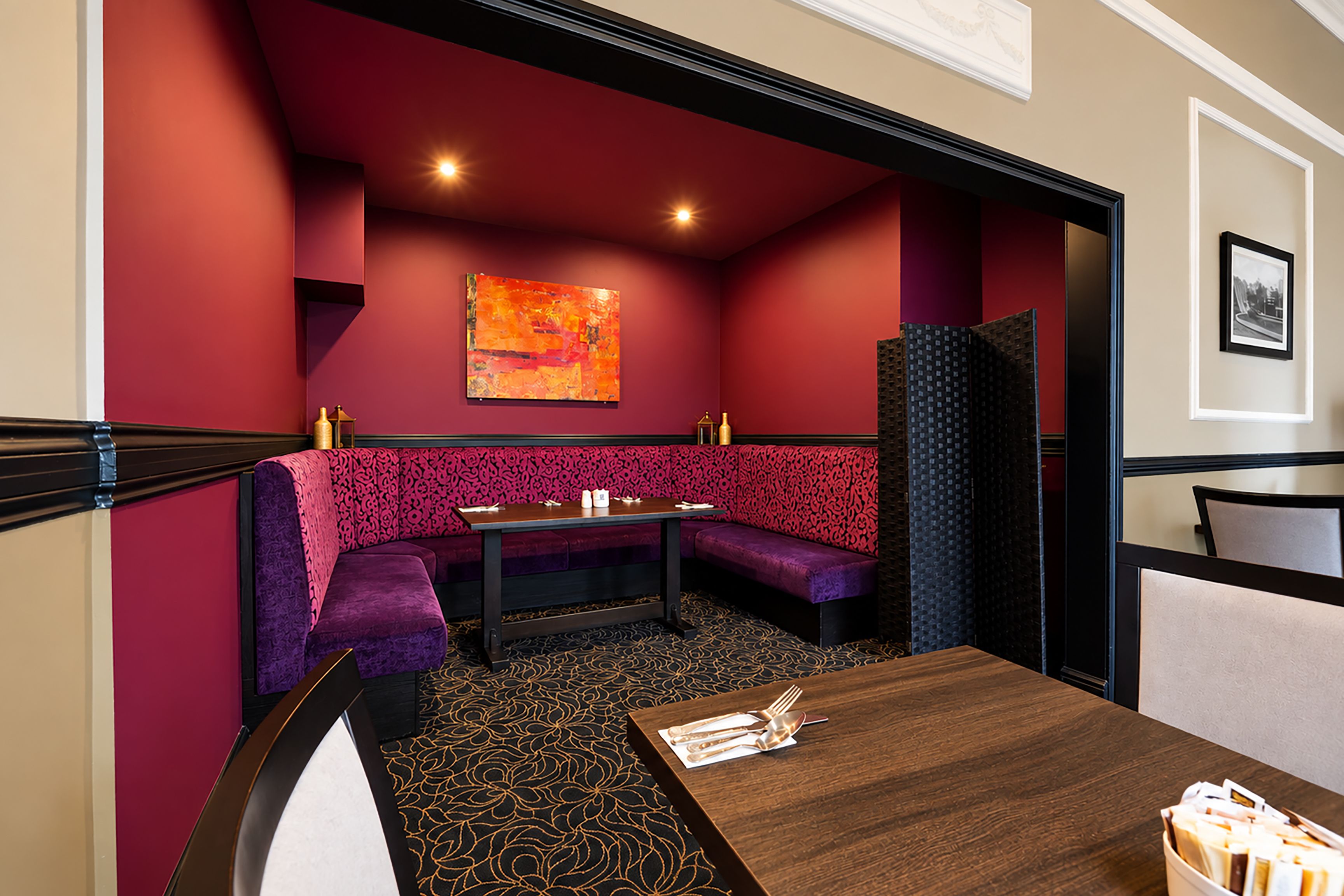 Una habitación con pared roja, alfombra estampada en negro y dorado, una mesa de madera con un menú, y un banco tapizado en color morado.