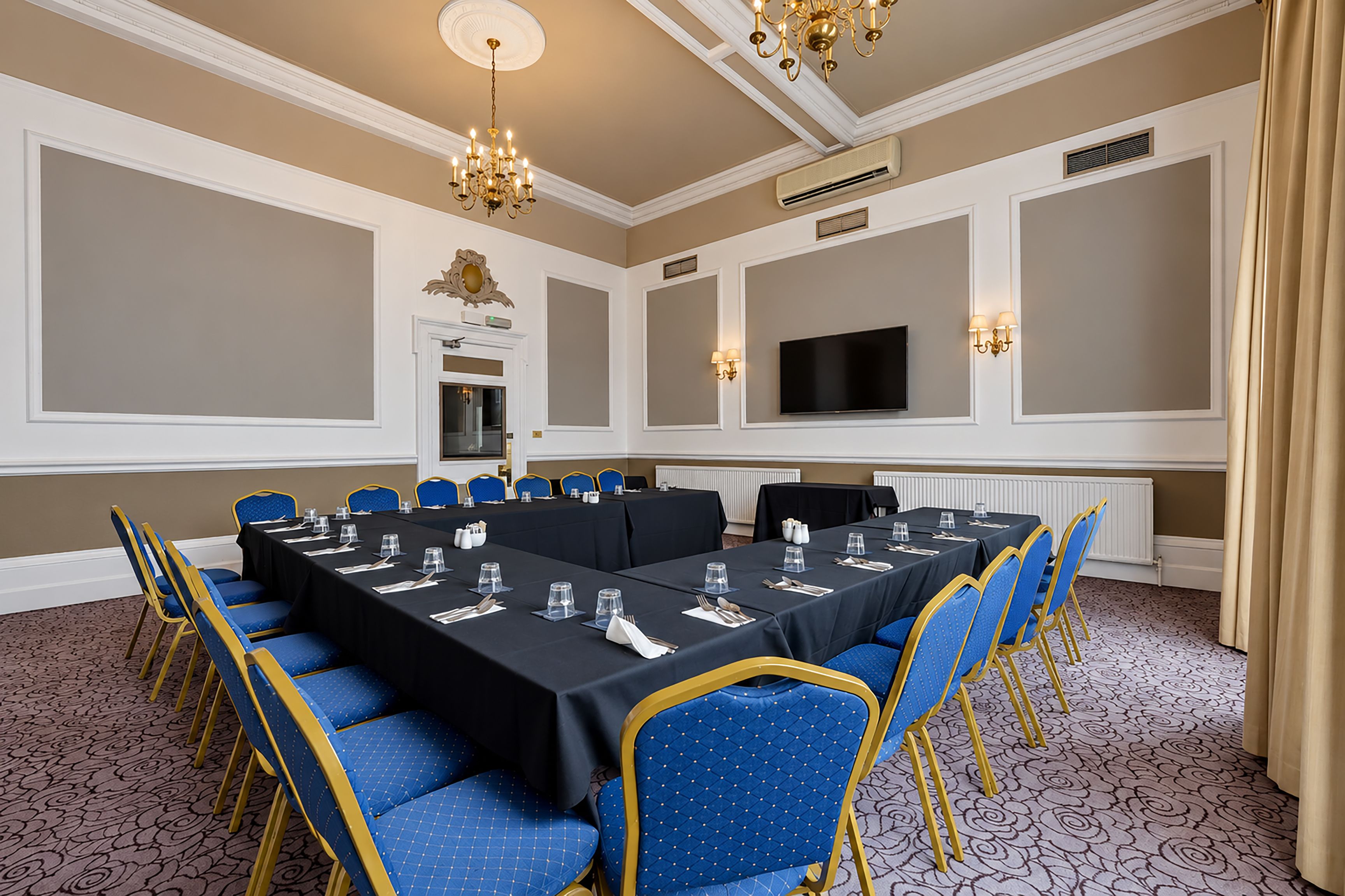 A conference room with a long table, blue chairs, a chandelier, and a flat-screen TV on the wall.
