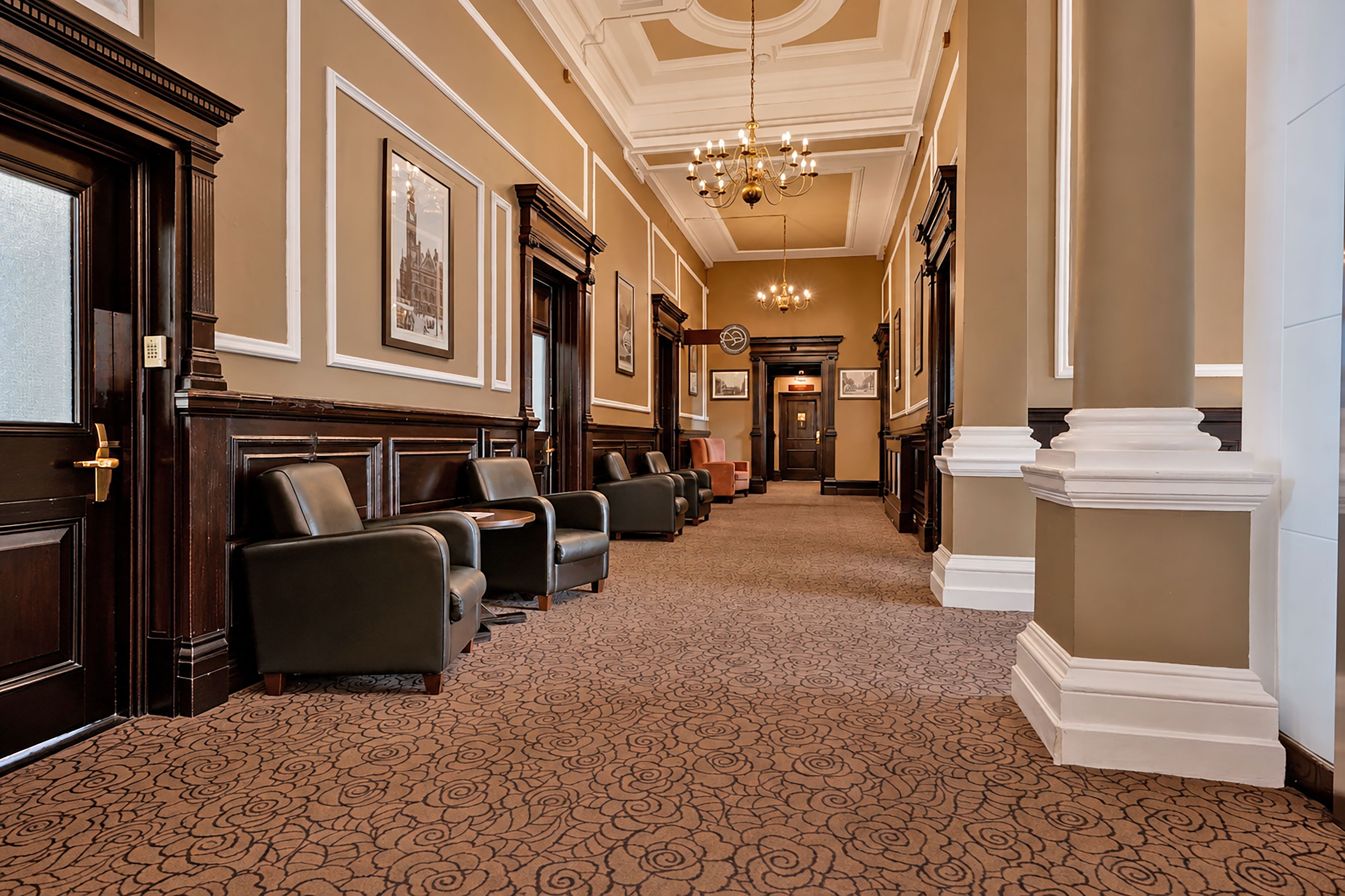 A hotel lobby with a carpeted floor, seating area with leather chairs, a chandelier, and framed pictures on the walls.