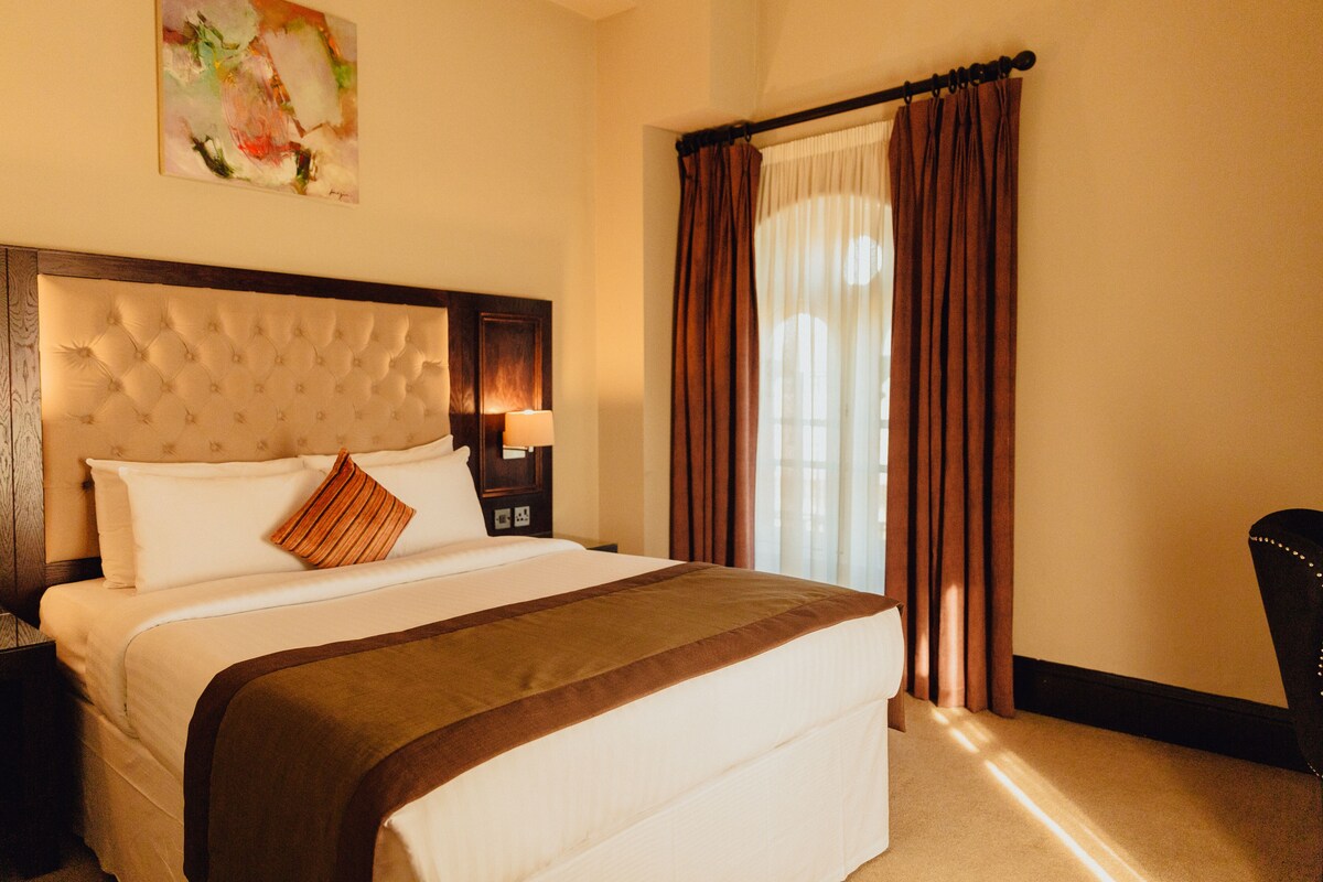 A hotel room with a large bed, a headboard, a nightstand with a lamp, and a window with curtains.