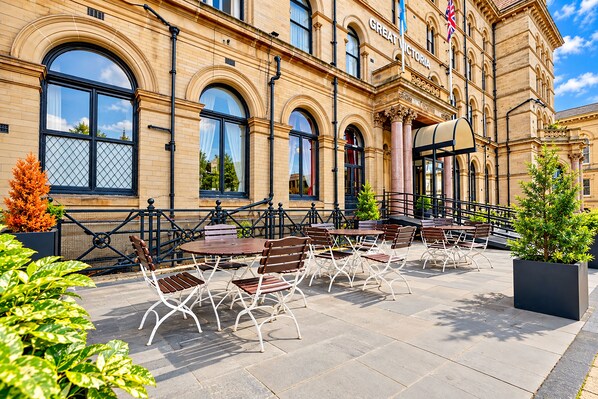An outdoor seating area with tables and chairs, a brick building with arched windows, and a flag. - The Victoria Hotel, Bradford by Sunday (Bradford)