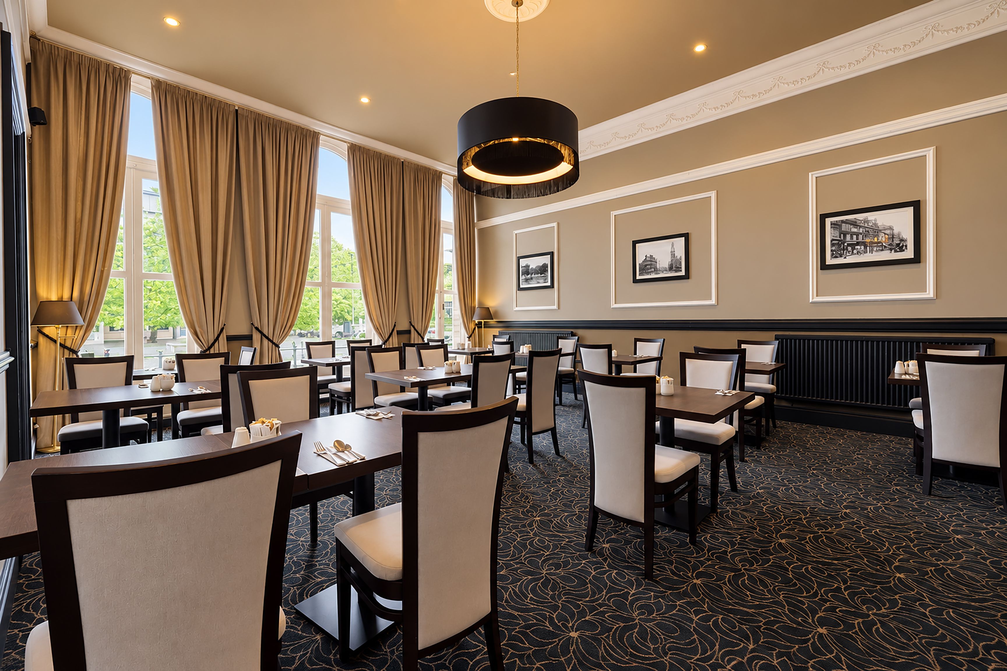A hotel dining area with multiple tables and chairs, large windows with curtains, and framed pictures on the walls.