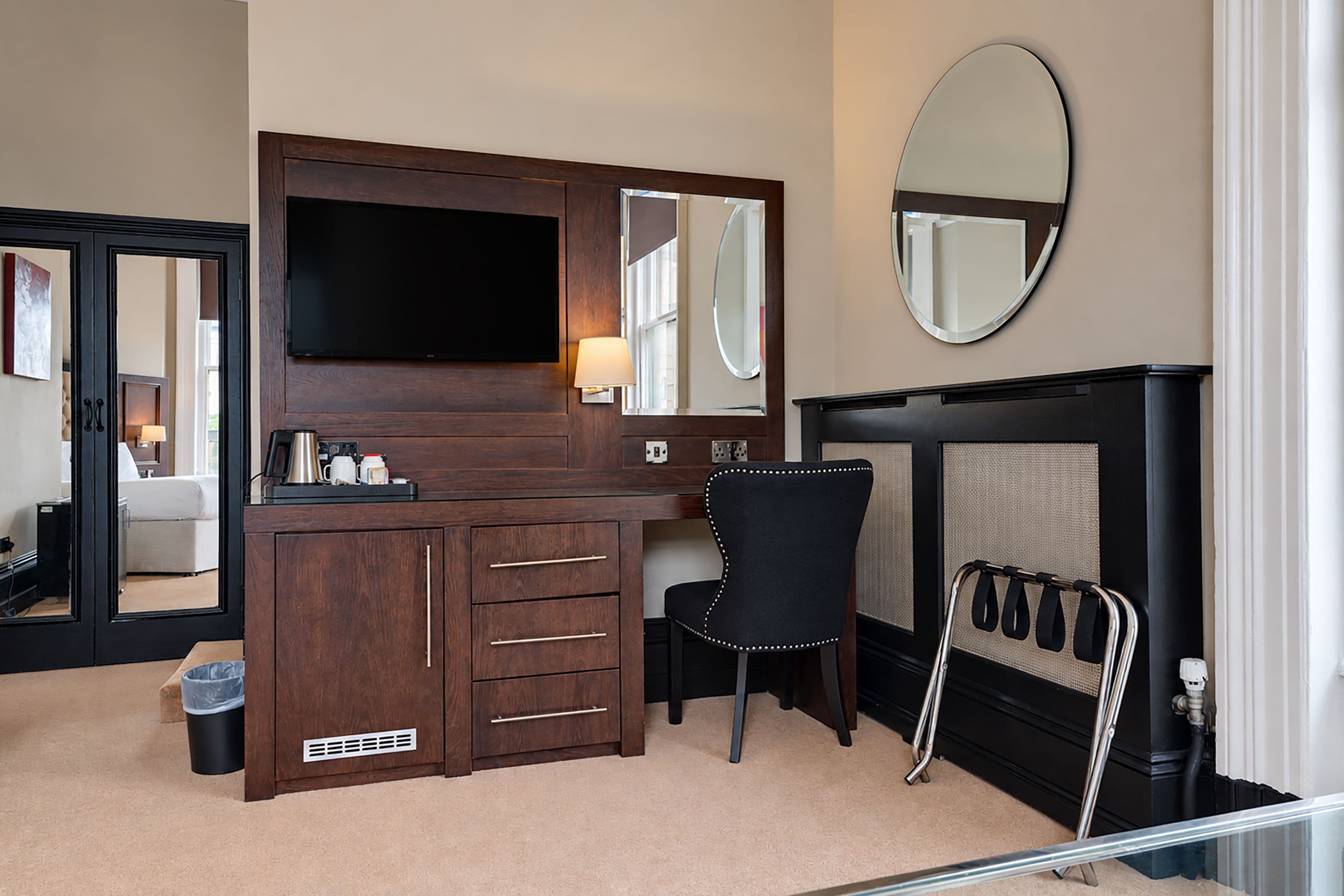 A hotel room with a wooden dresser, a flat-screen TV, a black chair, a fireplace, and a mirror on the wall.