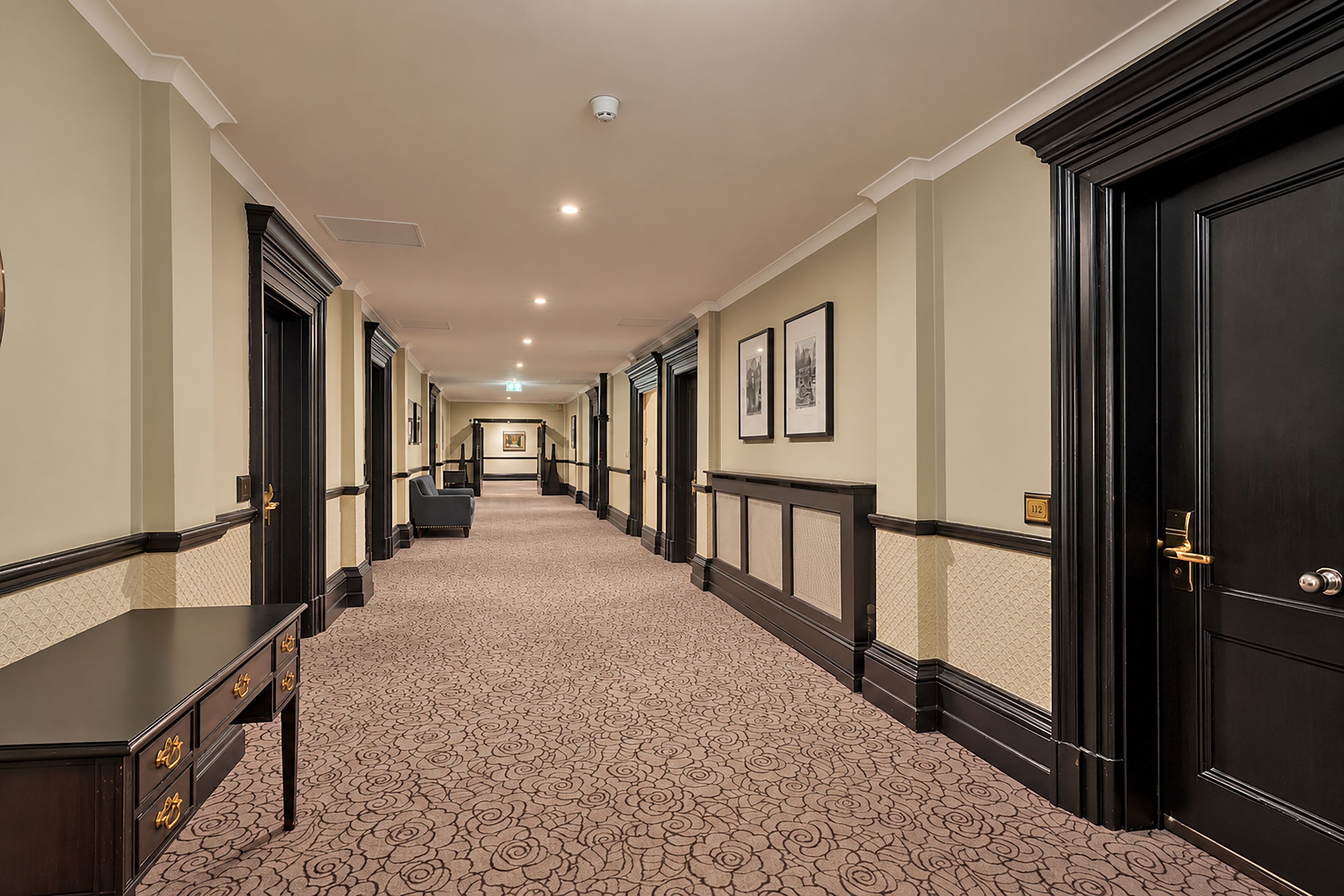A hotel corridor with a patterned carpet, dark wooden doors, and framed pictures on the walls.