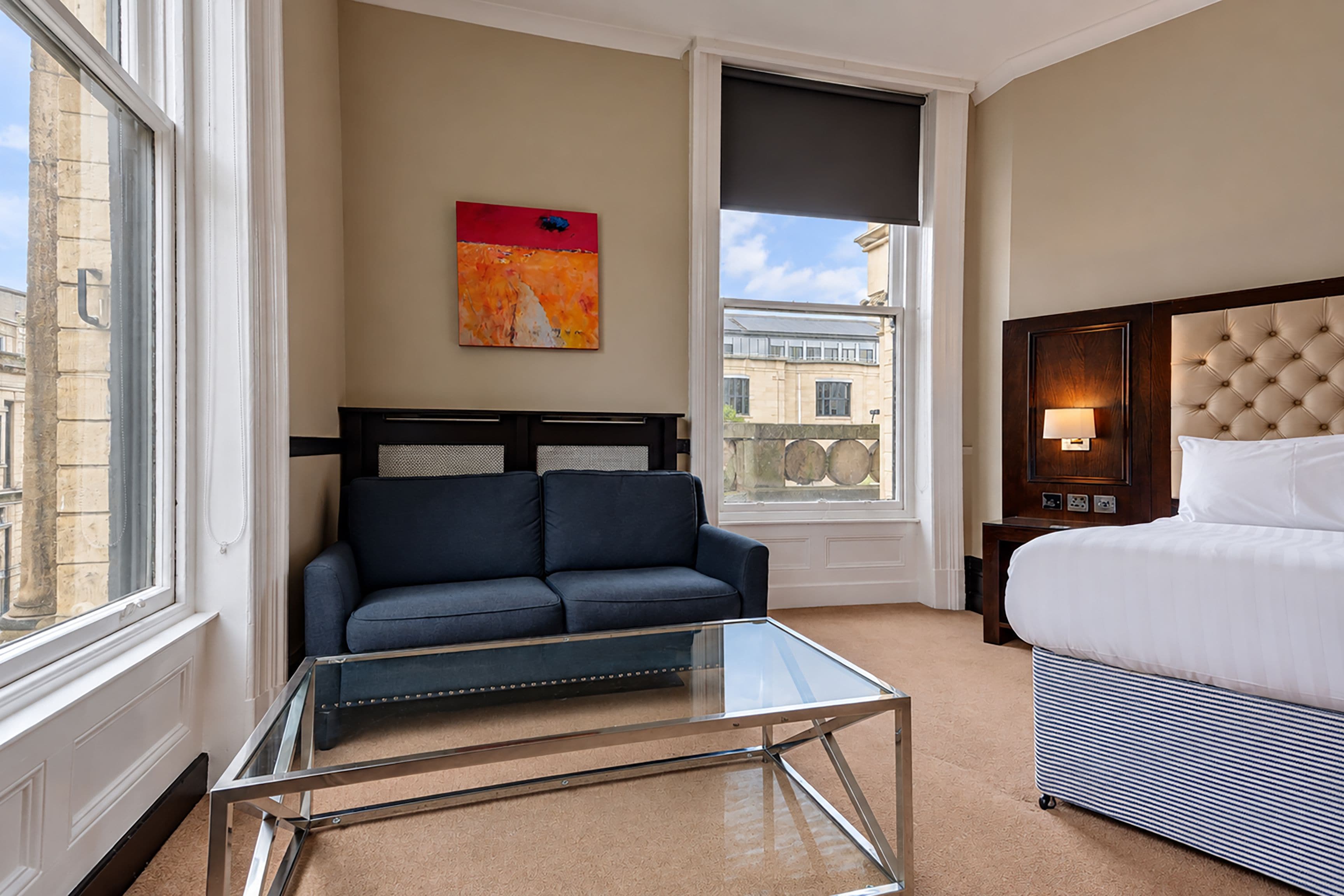 Una habitación de hotel con una cama, un sofá, una mesita de vidrio, un cuadro en la pared y vista a la ciudad por la ventana.