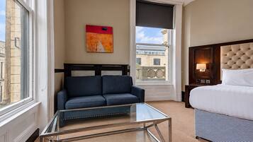 Quarto de hotel com cama, sofá, mesa de centro de vidro, pintura na parede e vista da cidade pela janela.