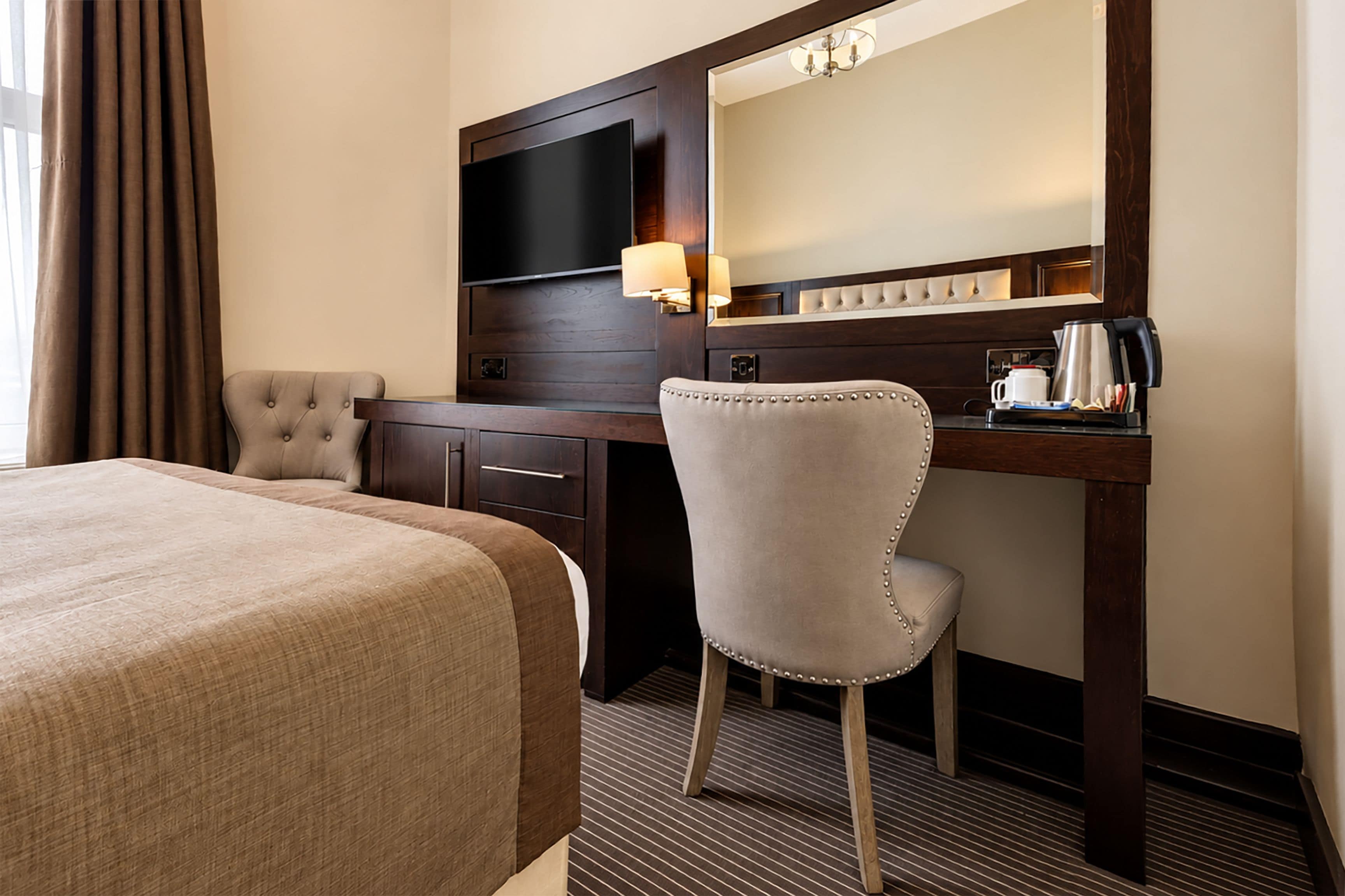 Ett hotellrum med en stor säng, ett skrivbord med en stol, en platt-TV, en spegel och en kaffebryggare.