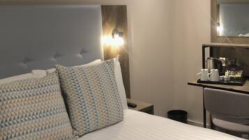 A hotel room with a large bed, a bedside table with a lamp, a small desk with a chair, and a mirror on the wall.