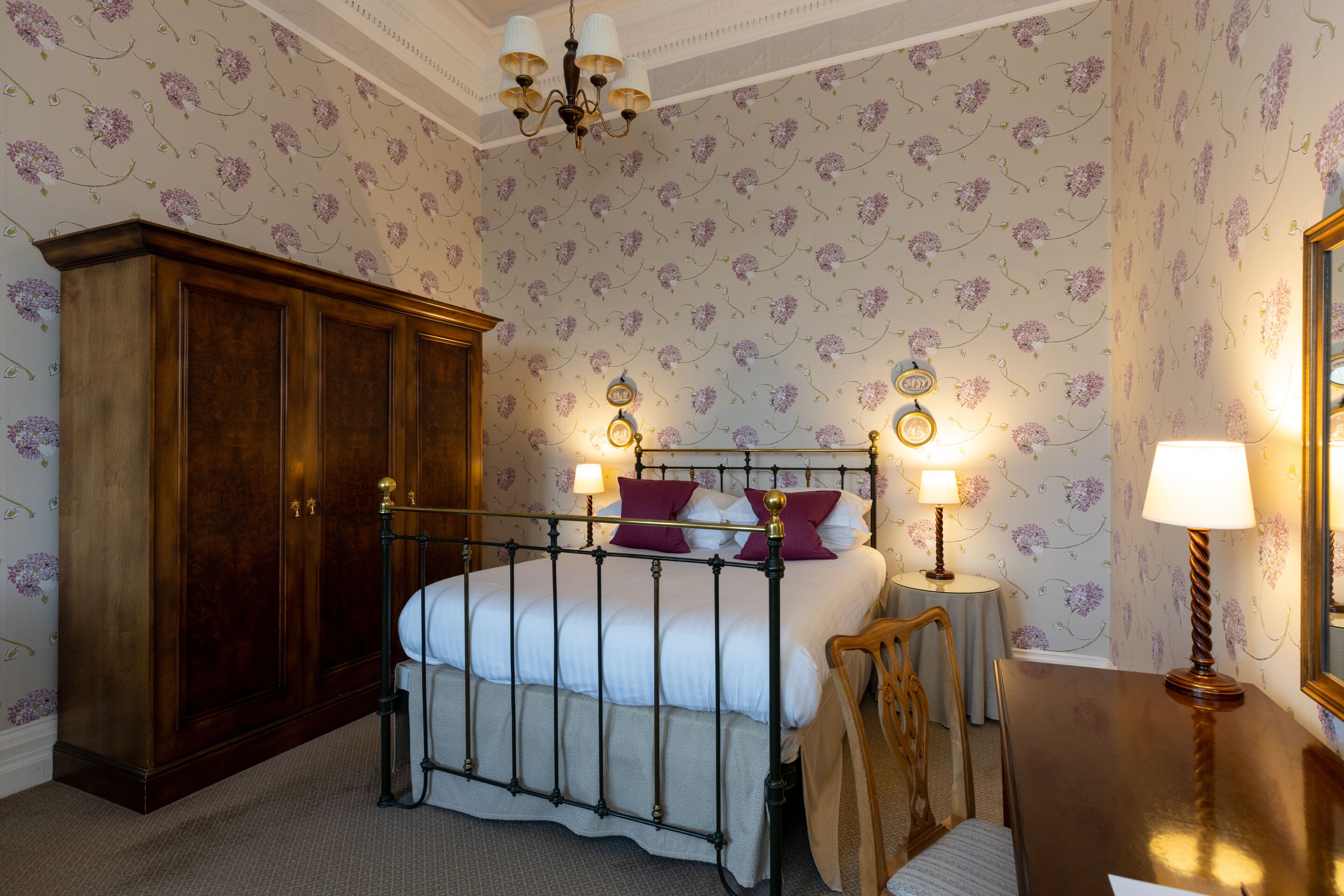 A bedroom with a four-poster bed, a wooden wardrobe, a round table with a lamp, and floral wallpaper.