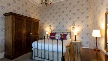 A bedroom with a four-poster bed, a wooden wardrobe, a round table with a lamp, and floral wallpaper.