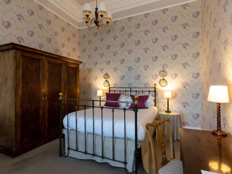 A bedroom with a four-poster bed, a wooden wardrobe, a round table with a lamp, and floral wallpaper.
