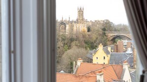 Vista desde una ventana con cortinas, que da a un edificio histórico y un puente, con una mesa y una silla en primer plano.