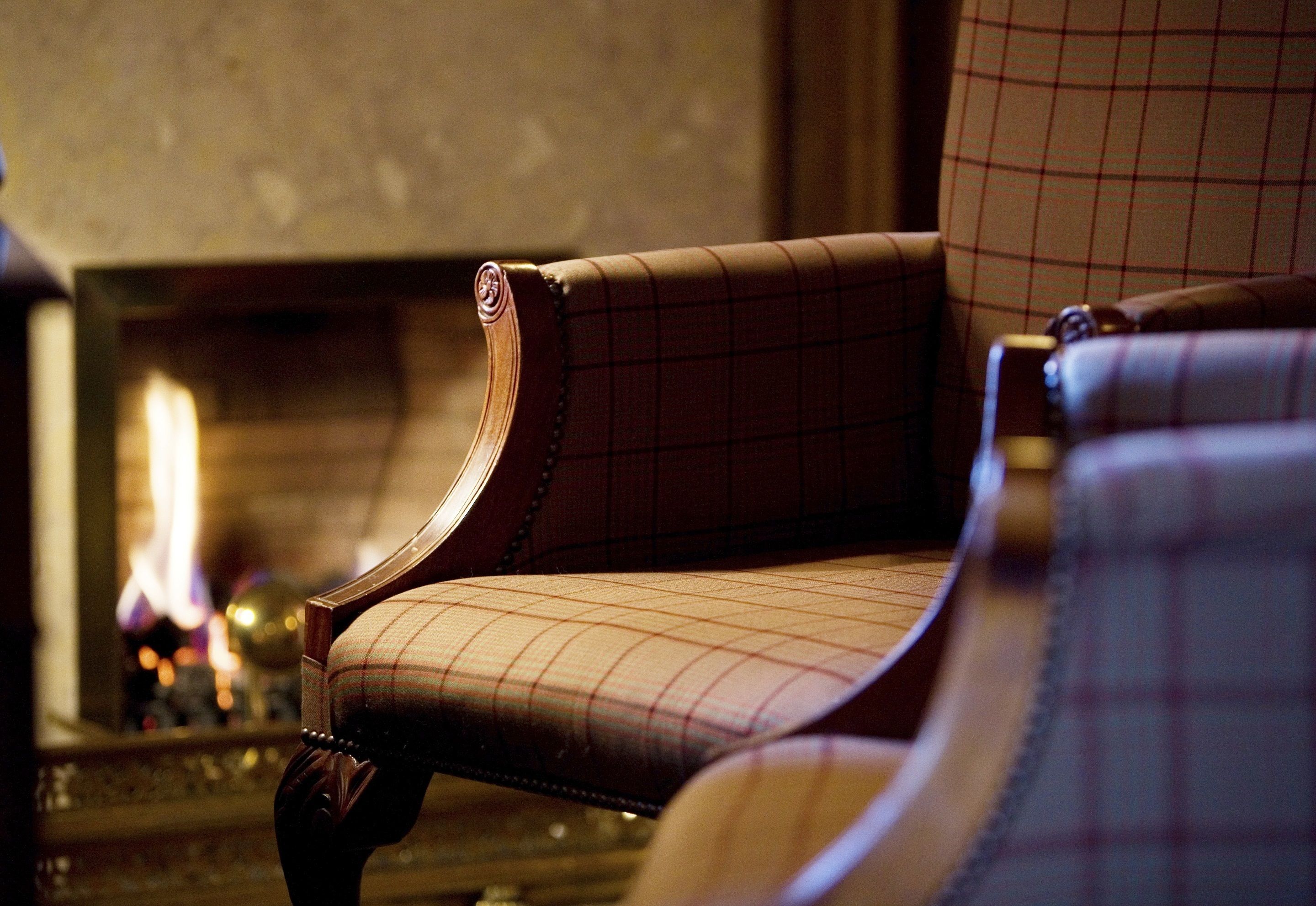 A close-up of a plaid-patterned armchair with wooden armrests, positioned in a warmly lit room with a fireplace in the background.
