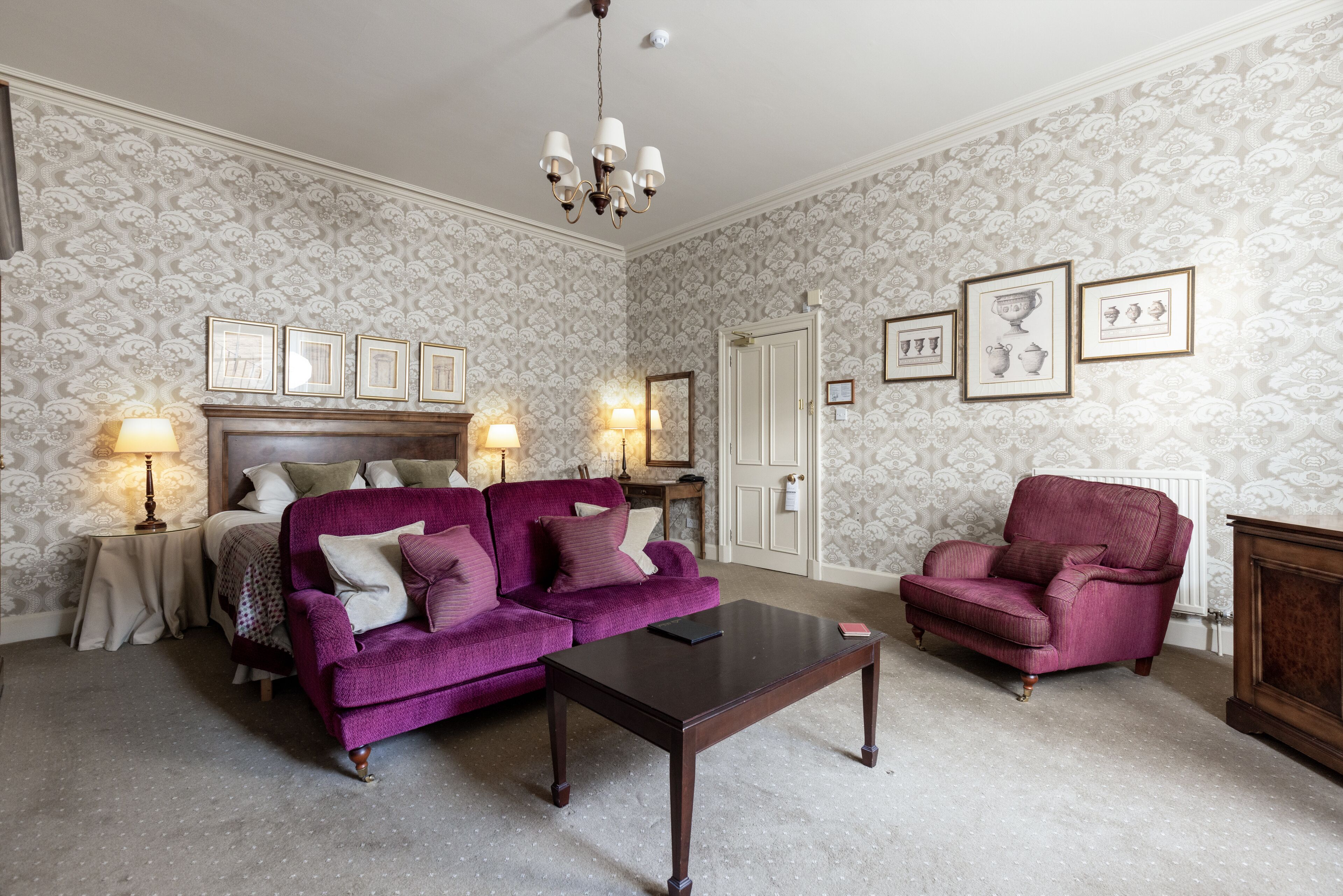 A room with a purple sofa, a wooden coffee table, a red armchair, a bed with a headboard, and a wall with framed pictures.