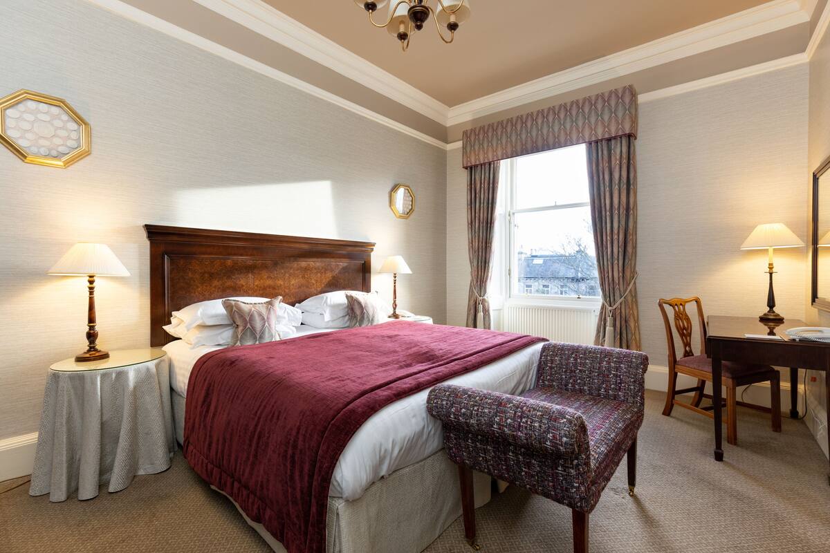 A hotel room with a large bed, two bedside tables with lamps, a chair, a wooden desk, and a window with curtains.