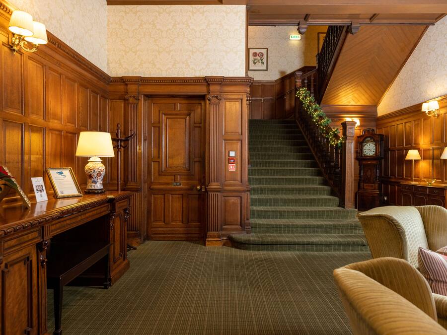 A grand lobby with a wooden reception desk, a staircase with a green carpet, and a cozy seating area with striped cushions.
