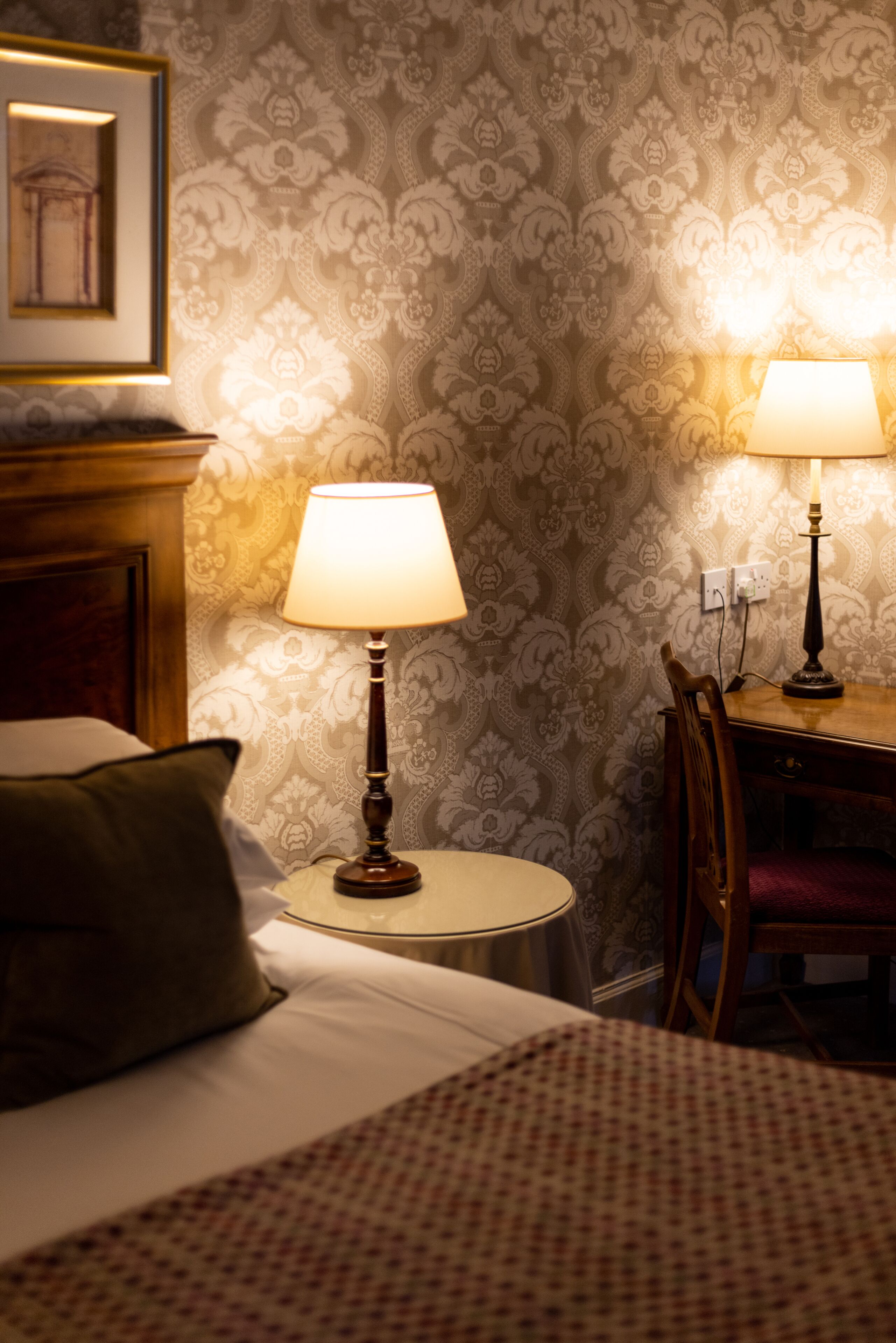 A hotel room with a bed, two bedside lamps, a wooden nightstand, a chair, and a patterned wallpaper.