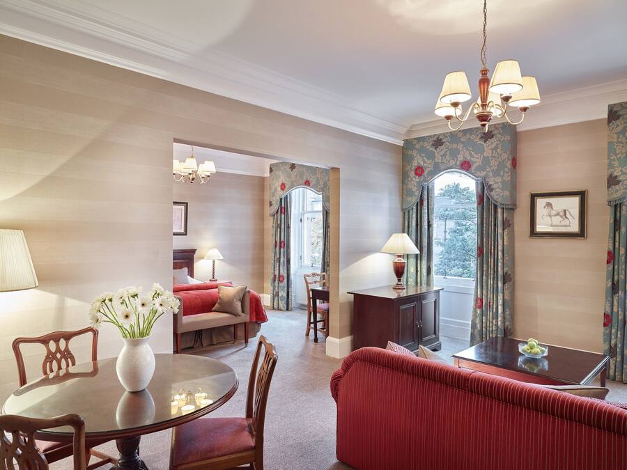 Ein Hotelzimmer mit Bett, einer Sitzecke mit Sofa und Sesseln, einem Esstisch mit Stühlen, einer Kronleuchter und einem Vorhangfenster.