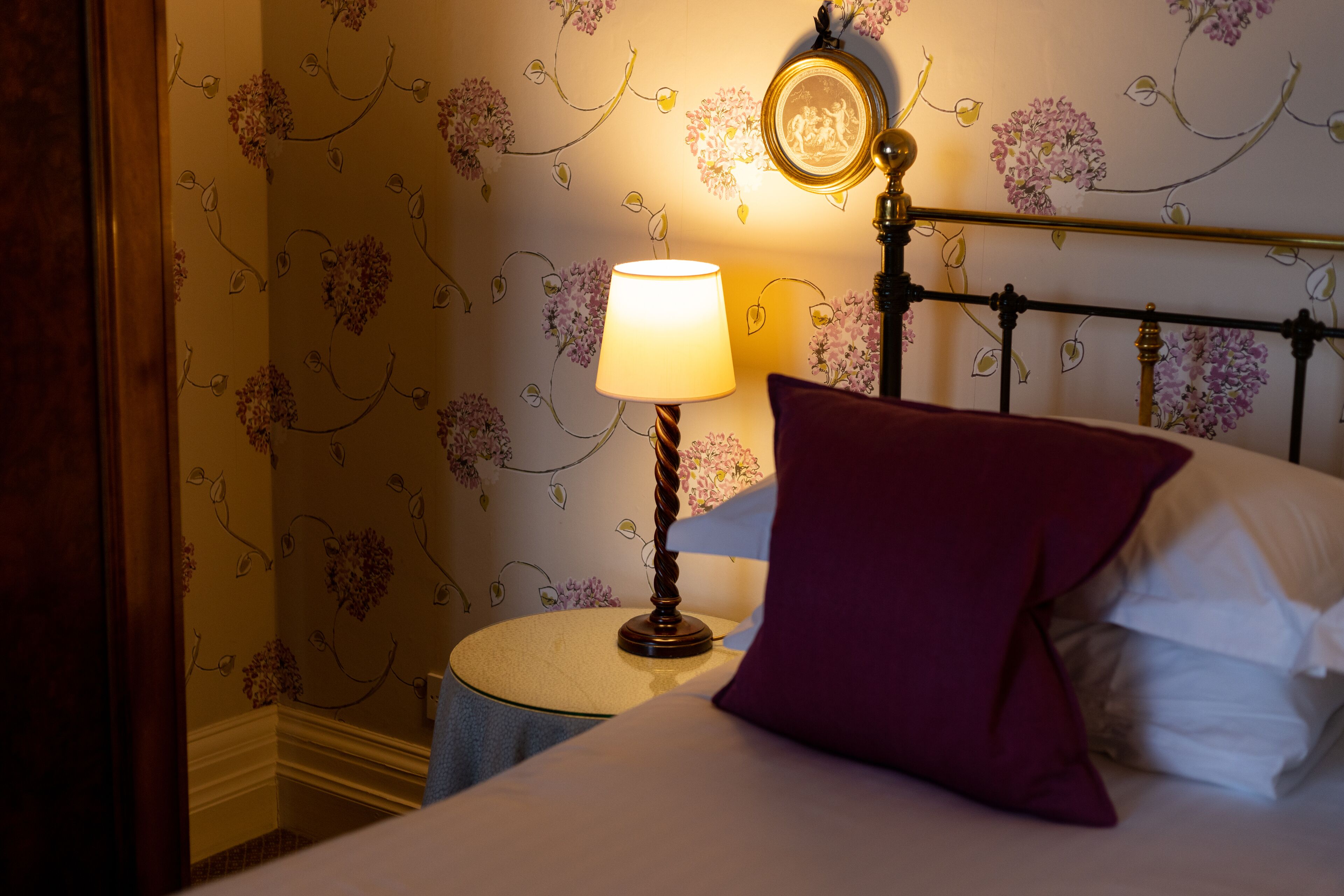 Una cama bien hecha con un cojín morado, una lámpara de noche y un espejo montado en la pared.