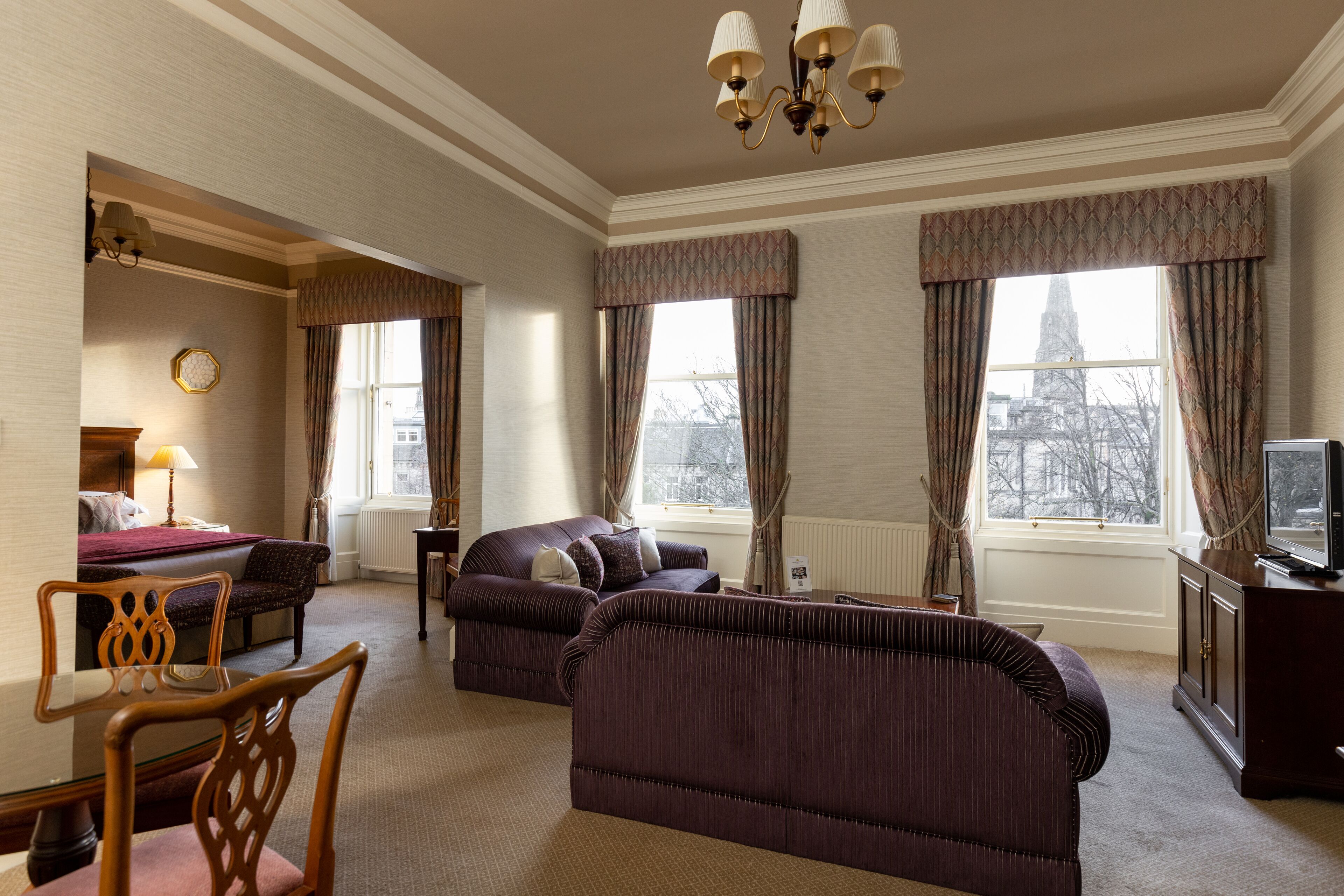 Une chambre d’hôtel comprenant un lit, deux canapés, une table à manger avec des chaises, un téléviseur et de grandes fenêtres avec des rideaux.