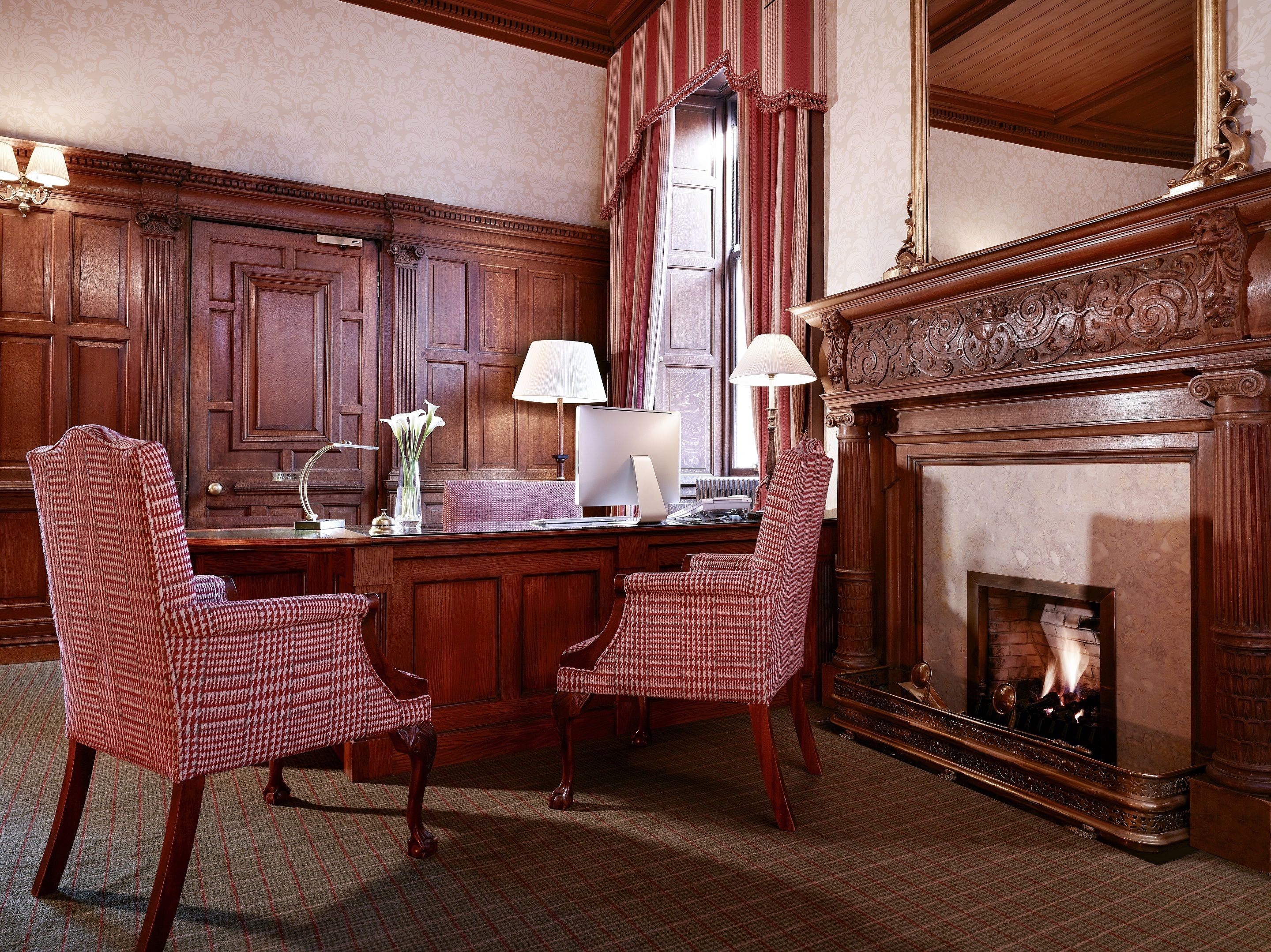 A traditional room with a wooden desk, two chairs, a fireplace, and a large mirror.