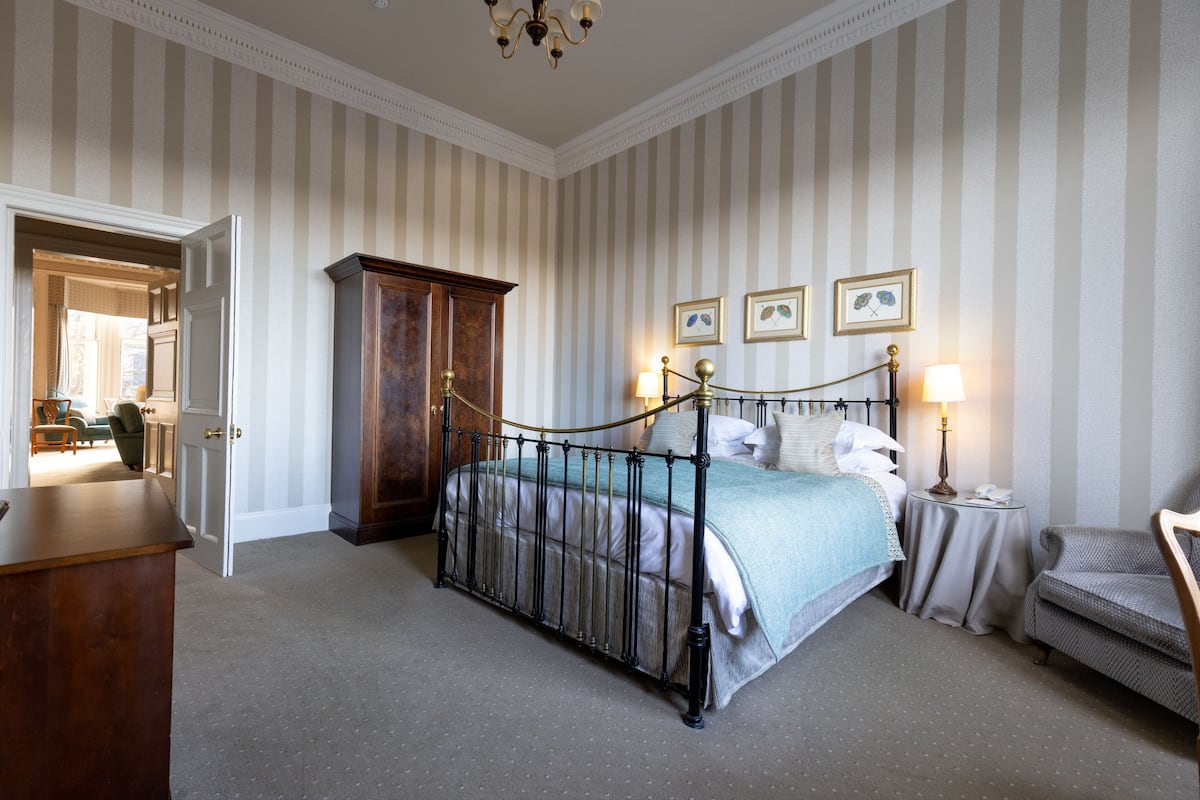 A bedroom with a four-poster bed, a wooden wardrobe, a small bedside table with a lamp, and a chair.