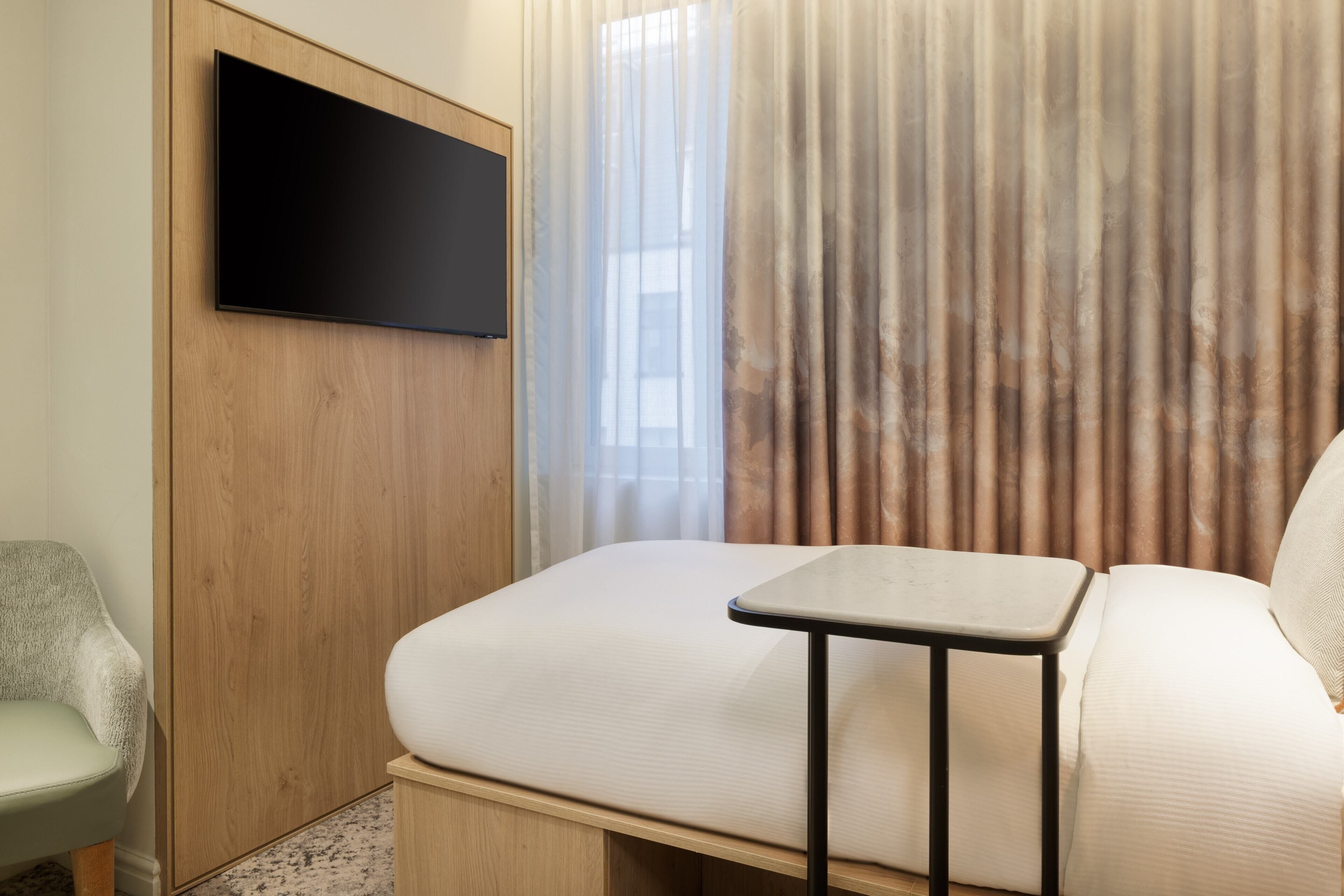 Une chambre moderne équipée d’un lit, d’une petite table, d’une chaise et d’un téléviseur fixé sur un panneau en bois.