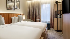 A hotel room with two beds, a small round table, a chair, a TV, and a wardrobe. - The Memoir Club (London)