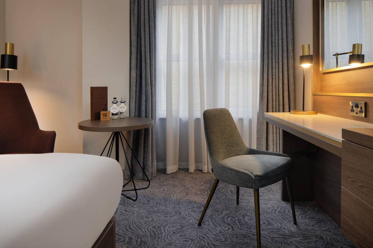 A hotel room with a bed, a desk, a chair, a round table with a lamp, and a mirror.