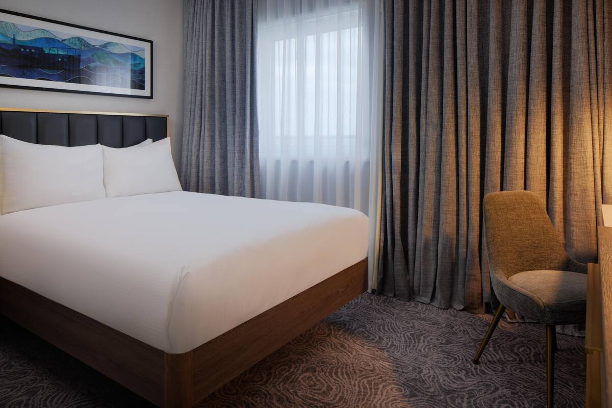 A hotel room with a large bed, a chair, a painting on the wall, and a window with curtains.