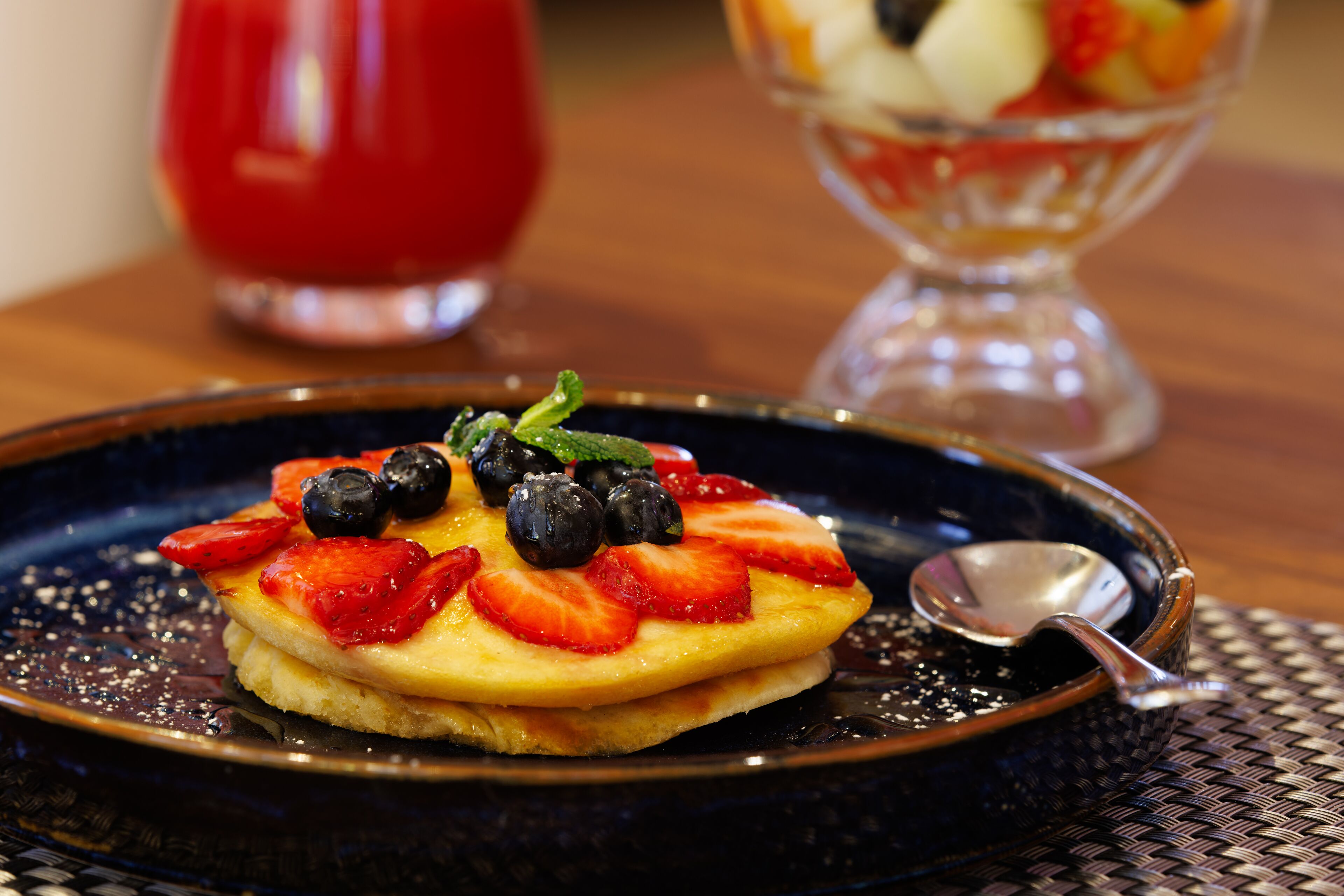 A stack of pancakes topped with strawberries and blueberries on a dark plate with a spoon.