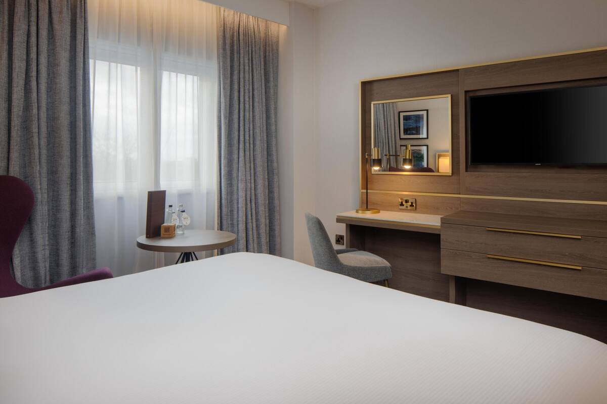 A hotel room with a bed, a desk, a chair, a TV, and a small table with bottles.