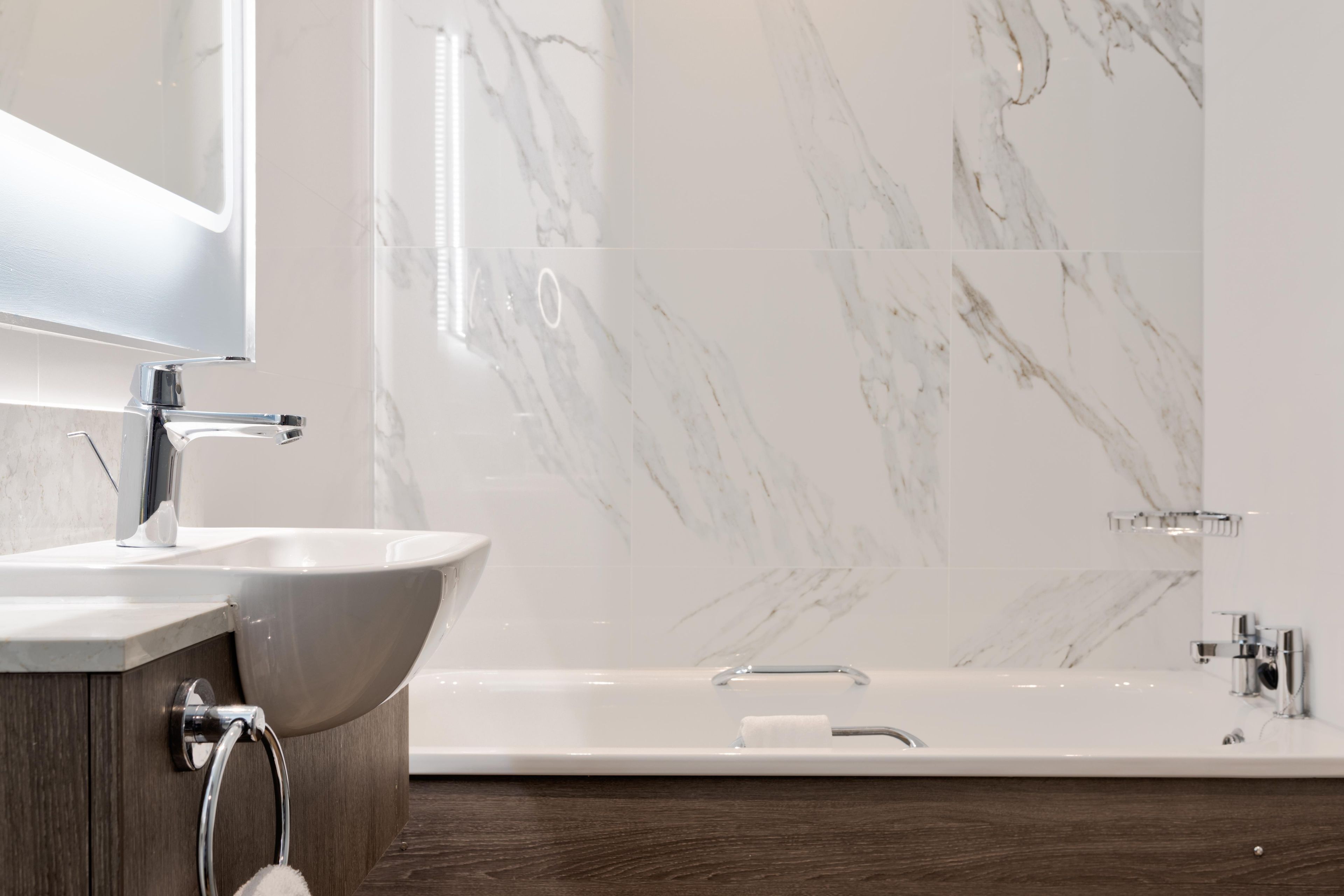 A modern bathroom with a white sink, a mirror, a bathtub, and a marble wall.