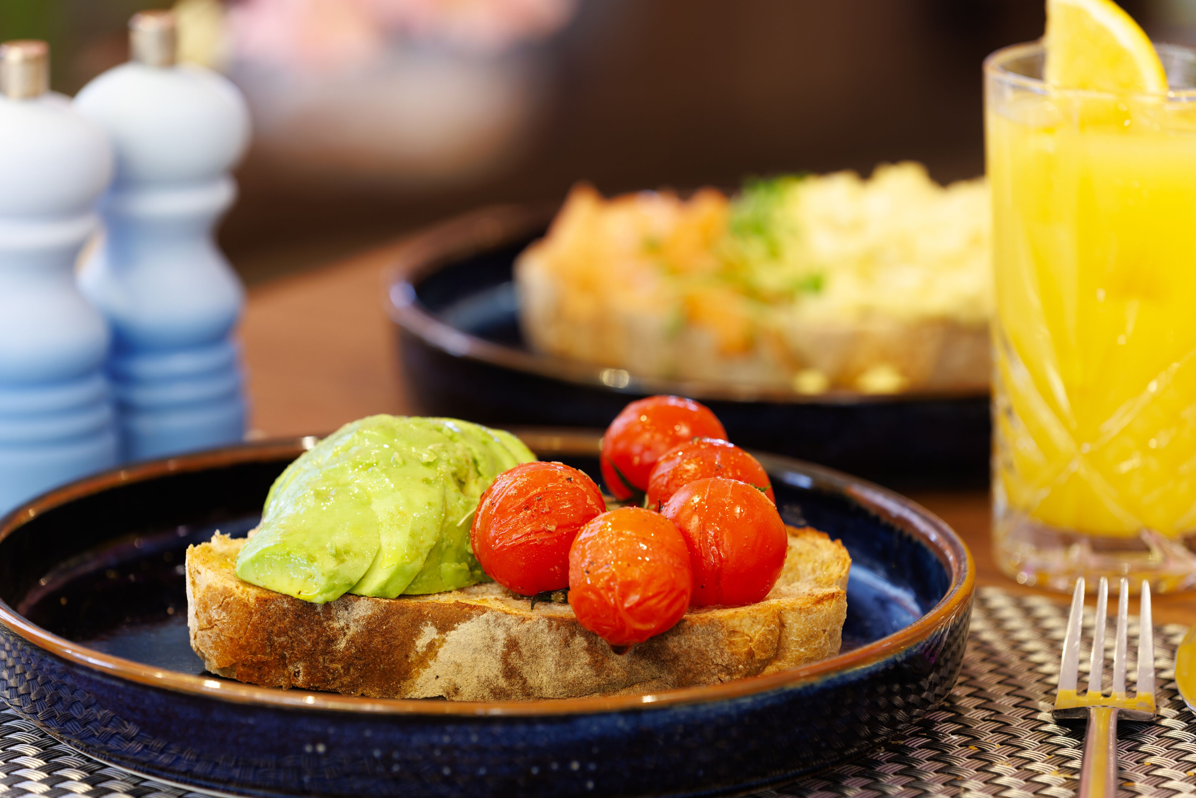 Une assiette de petit-déjeuner avec de l’avocat et des tomates cerises sur une tranche de pain grillé, un verre de jus d’orange et une fourchette posée sur un set de table en fibres tressées.