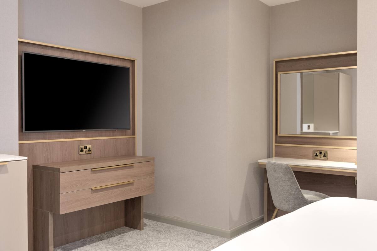 A hotel room with a flat-screen TV, a wooden dresser with drawers, a desk with a chair, and a large mirror.