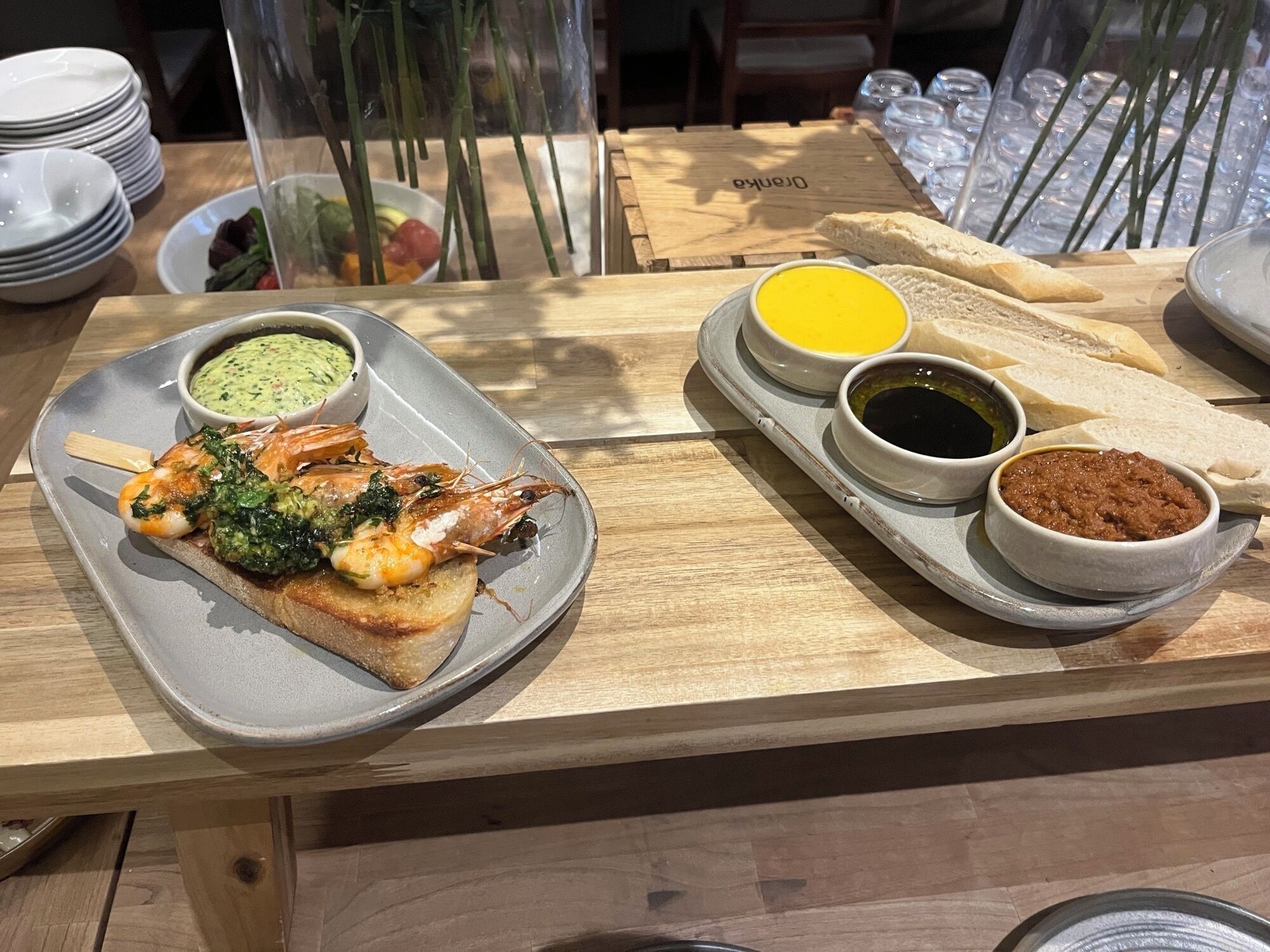A dining table with a plate of grilled shrimp, a slice of bread, and three small bowls of condiments.