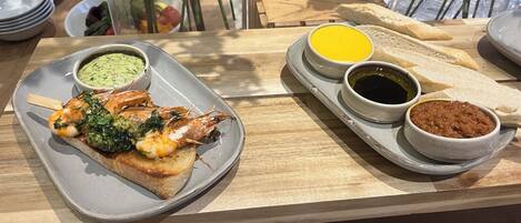 Een eettafel met een bord gegrilde garnalen, een sneetje brood en drie kleine schaaltjes met bijgerechten.