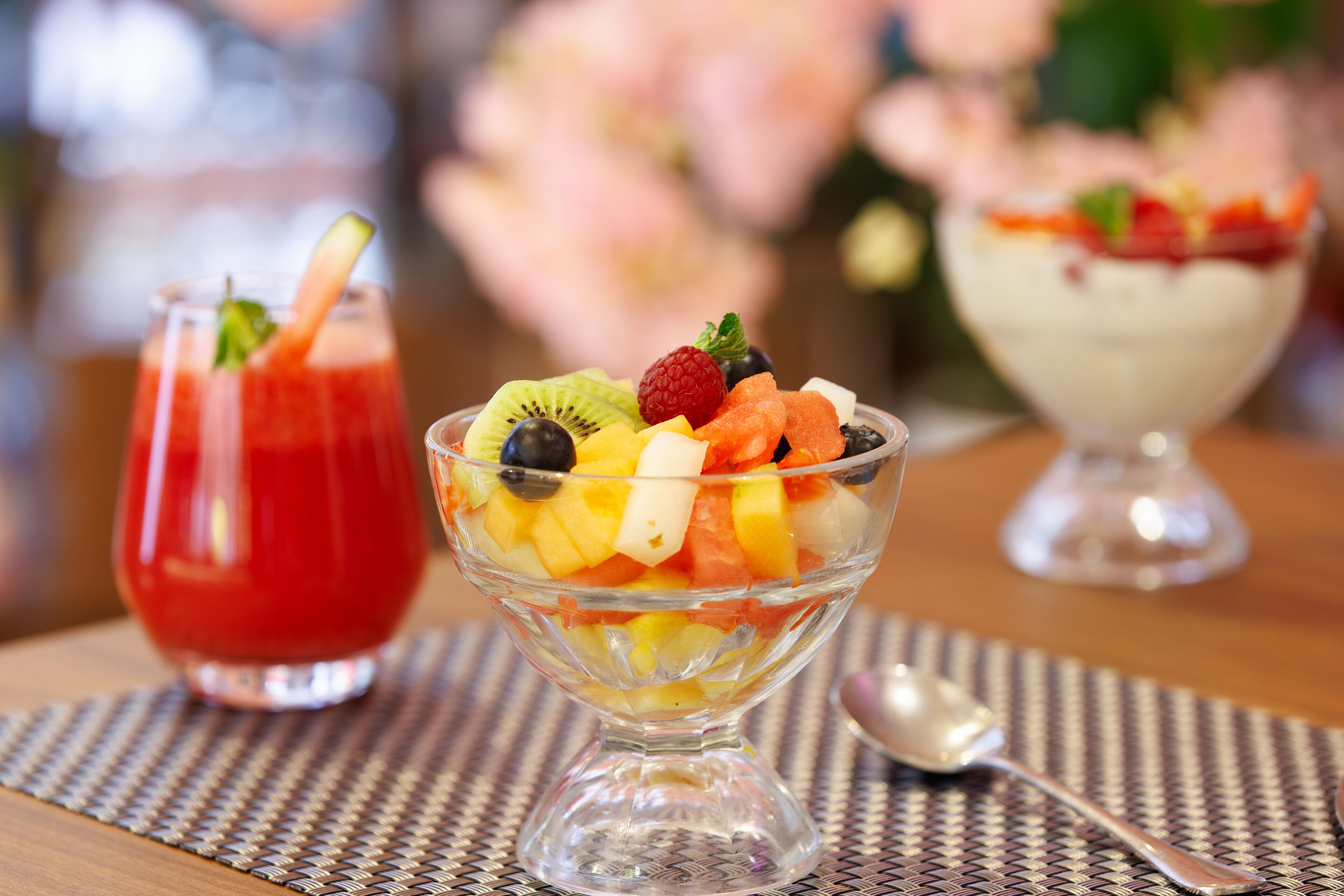 A dessert table with three bowls of fruit and a glass of red drink.