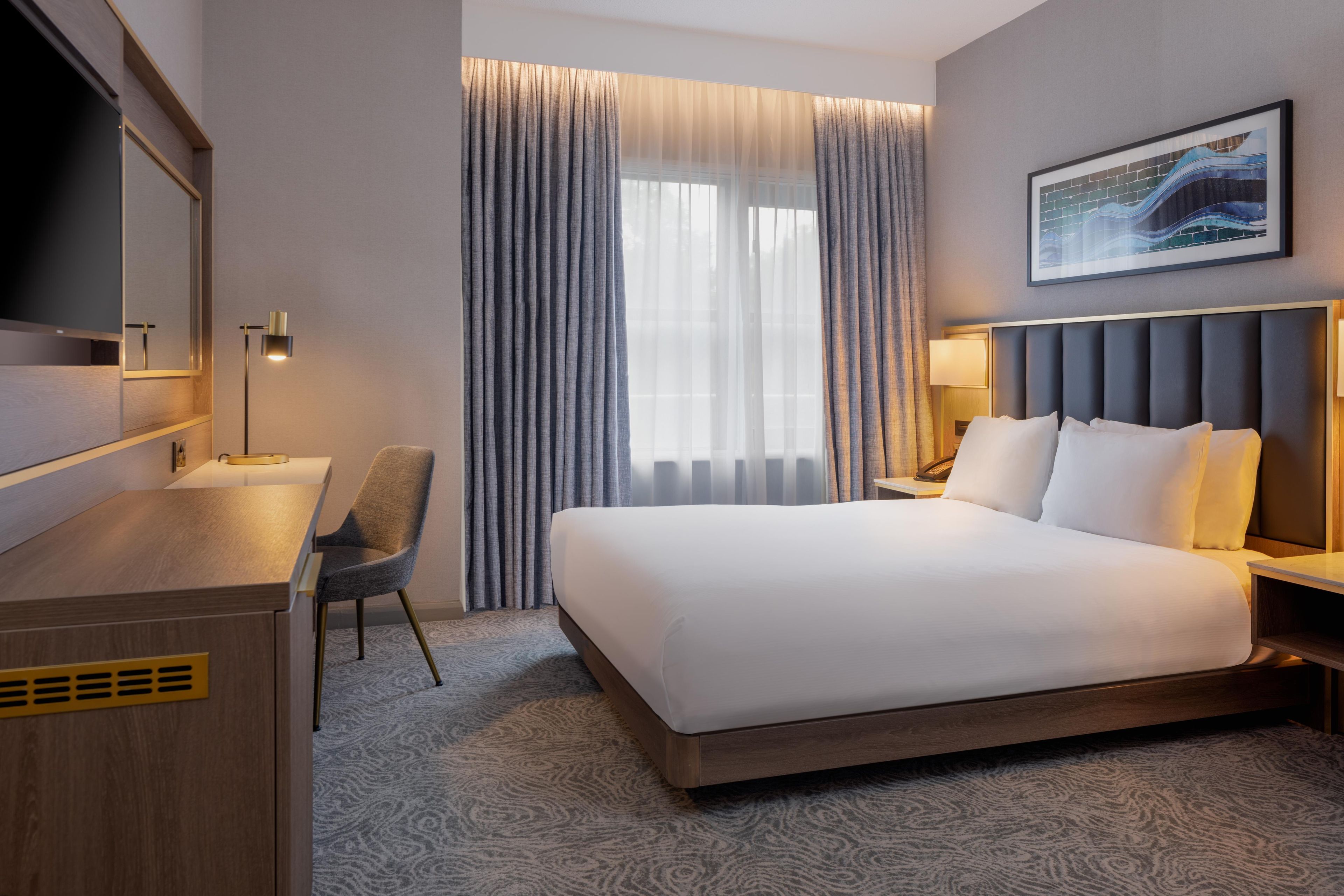 Een hotelkamer met een bed, een bureau, een stoel, een nachtkastje, een lamp, een schilderij en een raam met gordijnen.