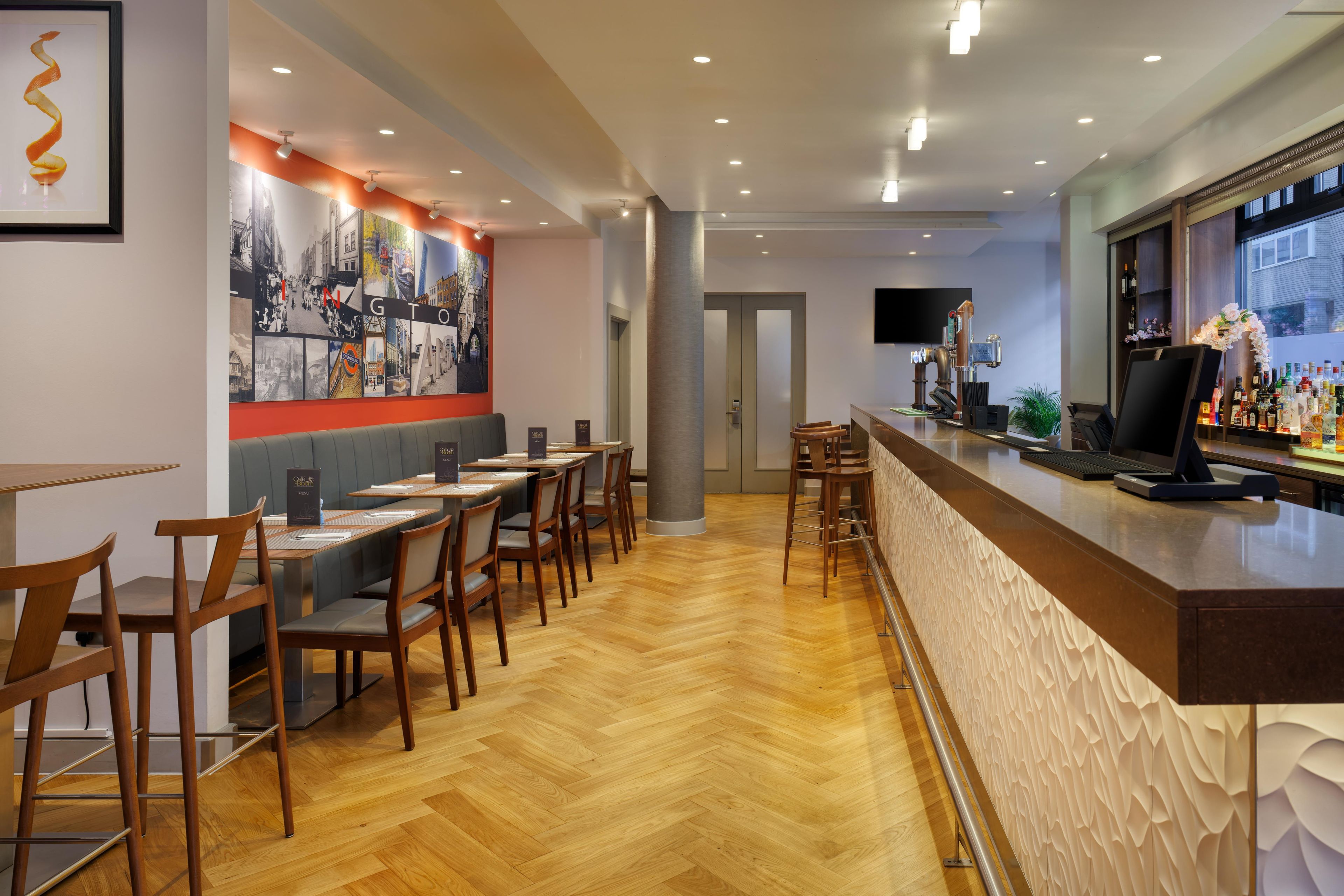 Angolo bar con sedie e tavoli di legno, un bancone bar con diverse bottiglie e un murale raffigurante un panorama cittadino.