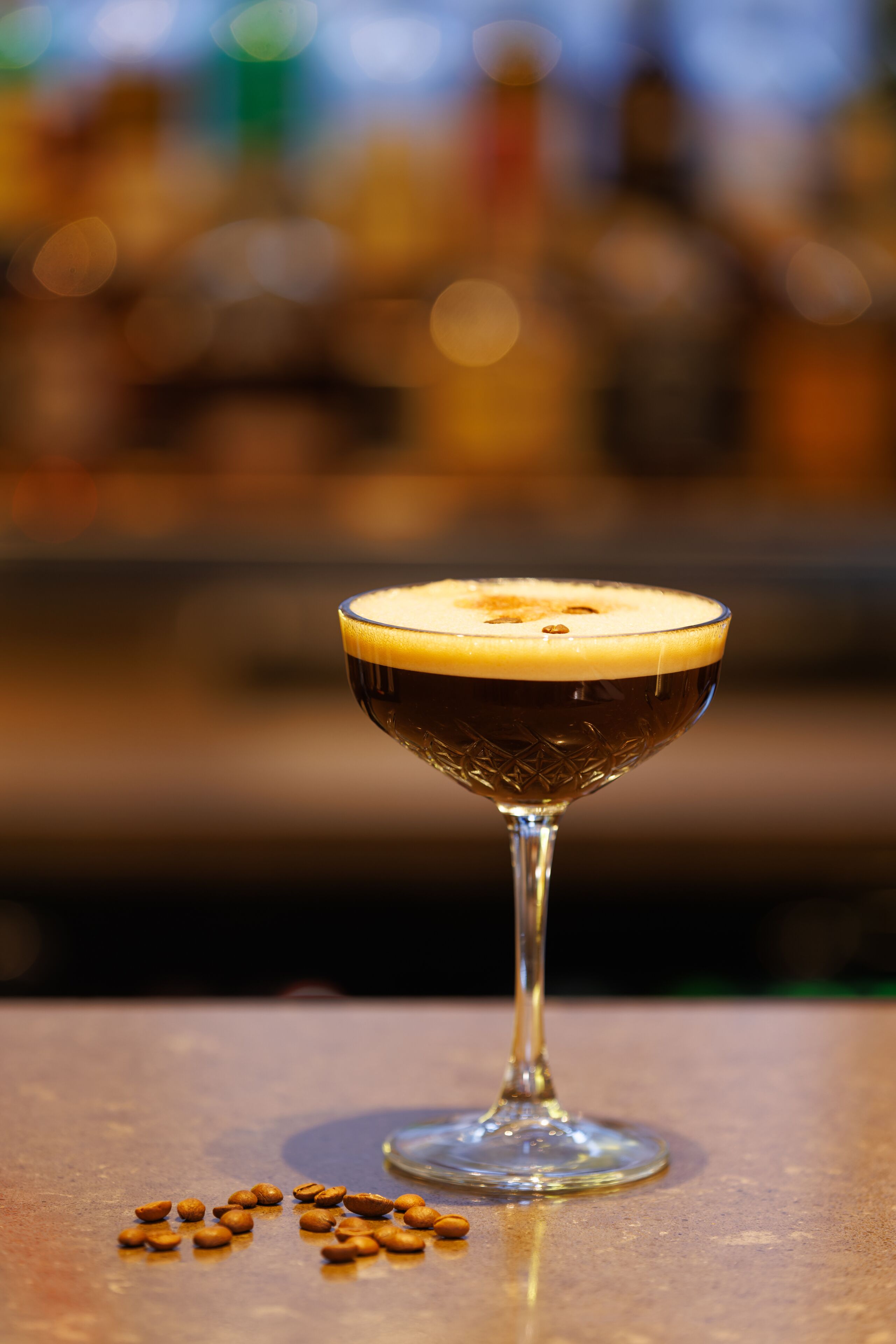 A cocktail in a stemmed glass with a frothy top, placed on a surface with scattered coffee beans.