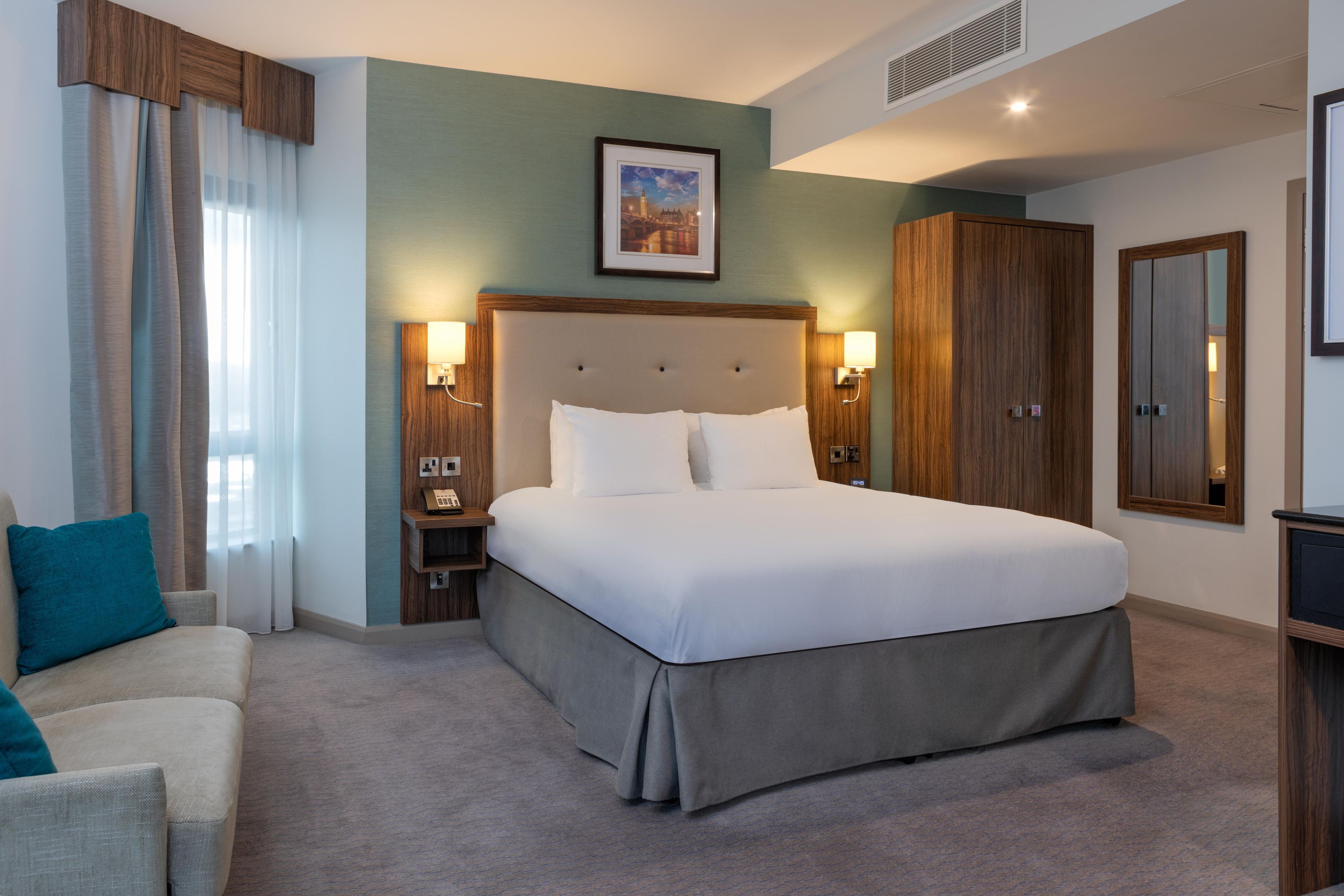 A hotel room with a large bed, a nightstand, a desk, a chair, and a wardrobe.